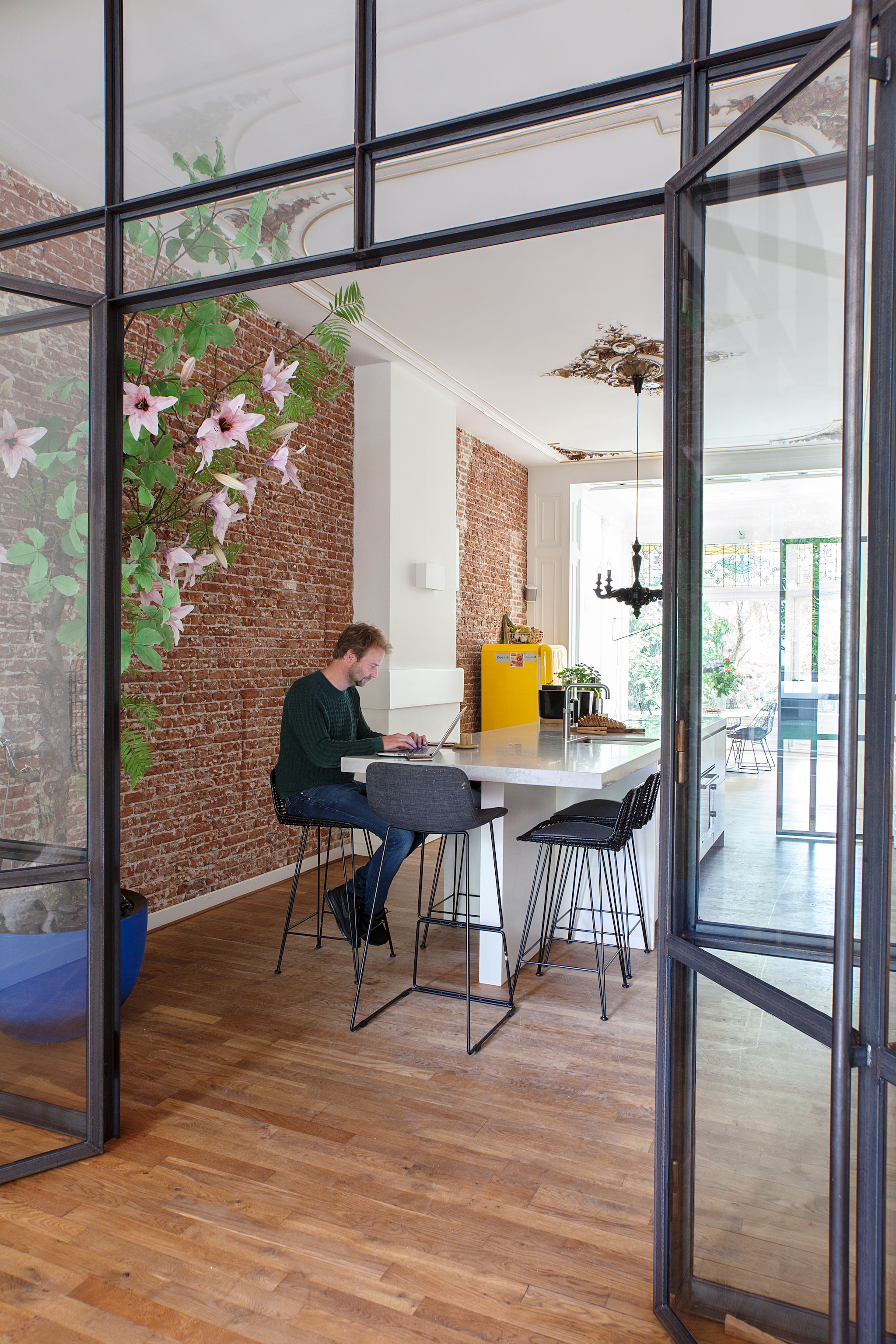 Keuken tussen zitkamer en oranjerie, gescheiden door stalen kozijnen, zelf ontworpen. Boom met kunstbloemen in blauwe pot ’s Zomers Bloemen. vtwonen binnenkijken special 12-2020 | fotografie Corine Hoge Bavel | styling Sheila Sletbeen via Kieveen