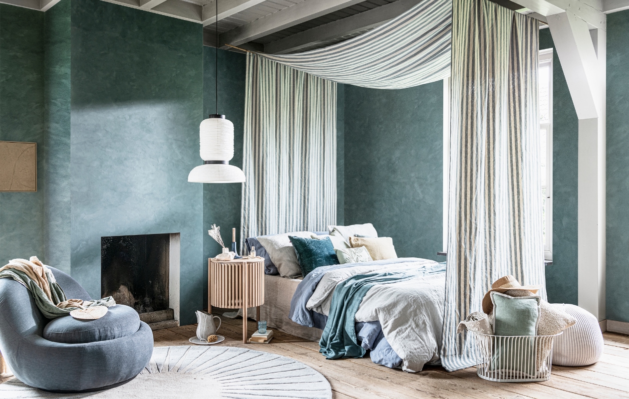 Kalkverf Blue Reef op de wand, per liter € 47,- (pure-original.nl). Schilderij met zandtextuur € 250,- (mooswillemsen.com). Fauteuil Babette, 84x93x73 cm (bxdxh) € 499,- (sofacompany.com). Wollen vloerkleed, 200 cm (Ø) € 399,- (made.com). Turkooizen-beige plaid € 139,-, poef € 430,- (homestede.nl). Bijzettafel 44x64 cm (Øxh) € 969,- (bolia.com). Hanglamp € 191,- (vantil.nl). Hemel gemaakt van stof Endless Summer van Barbara Osorio, 310 cm breed, per meter € 139,-, turkooizen velours kussen van William Yeoward € 98,-, turkooizen linnen plaid gemaakt van stof Canezza van Designers Guild, 297 cm breed, per meter € 94,-, opgerolde plaid van stof Theia van Nobilis, per meter € 238,- (wva.nl). Linnen laken in kleur Naturel, vanaf € 139,95, linnen dekbedovertrek in kleur Dusk Blue en Misty Green, vanaf € 199,95 (yumeko.com). Zachtgroen kussen met geborduurde bladeren € 22,99, zandkleurig kussen € 17,99, blauw linnen kussen € 19,99, zachtgroen kussen met witte bies € 22,99, gebroken wit kussen € 19,99, hoed € 22,99 (zarahome.com). Marmeren bord € 39,99, keramieken karaf € 29,99 (sissy-boy.com). Gebroken witte keramieken vaas € 36,- (serax.com). Houten kandelaar € 69,90 (letoile.store). Lichtblauw glas € 9,95 (anna-nina.nl). Metalen mand van Menu € 210,- (designtales.nl). Overig privébezit. vtwonen 08-2021 | styling Danielle Verheul | fotografie Sjoerd Eickmans