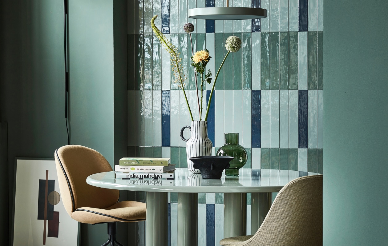 Glanzende wandtegels Marrakesh van vtwonen in kleuren Ocean Blue, Sea Green en Army Green, 6,5x40 cm, per m2 € 70,-, stalen wandlamp Arum van Ferm Living € 397,-, aluminium hanglamp Asteria van Umage € 205,95, groene glazen vaas Tummy van Urban Nature Culture € 25,95 (vtwonen.nl).Muurverf in Green Smoke No.47, per 2,5 liter € 97,- (farrow-ball.com). Eettafel Smalto van geëmailleerd staal van Knoll, 137x73 cm (Øxh) € 4888,50 (justhaasnoot.nl). Stoel links Beetle van Gubi in Divina 3, staal en textiel € 1249,-, stoel rechts Harbour Side Dining chair van Menu, kunststof en staal € 660,- (hellochair.nl). Zwart-witte kan € 24,99, zwarte schaal € 24,99 (hm.com). Poster Shapes of Colour 03 van Paper Collective, 50x70 cm € 49,-, eikenhouten lijst Quadrum, 50x70 cm € 64,95 (loods5.nl), meegeschilderd met de muur. vtwonen 10-2022 | styling Kim van Rossenberg | fotografie Peggy Janssen