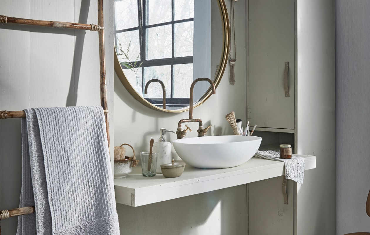 Grote ronde spiegel Ø 80 cm € 249,-, marmeren zeeppomp € 41,- (Nordal). Bamboe ladder € 54,05, vloerkleed 60x90 cm € 23,65 (Madam Stoltz). Witte wafeldoek € 49,95 (De Weldaad). Stoel Butterfly van Ercol € 625,- (De Machine- kamer). Berkenplex opbergmand 25x40x36 cm € 89,50 (Nordic New). Grijze handdoek 90x150 cm € 35,99(Zara Home). Waskom Girona, 57x40x15 cm € 381,- (Ton Scholten). Kraan € 75,- (Jan van IJken Oude Bouwmaterialen). Tadelakt zandkleurig potje met deksel € 19,95 (MaisonNL). Bureaulamp € 95,-(Vint. Amersfoort). Glas € 3,95, linnen doek in opbergzak € 75,-, witte opgerolde wafeldoek € 45,- (De Weldaad). Zandkleurig met bruine kom € 39,99, messing muurknop van Ferm Living € 20,-, geurkaars in bruin glazen potje € 16,- (Six and Sons). Rieten mand in kast vanaf € 9,99 (H&amp;M Home). Badborstel € 8,95 (Dille &amp; Kamille). De kast is geschilderd in de kleur Bone van Farrow &amp; Ball per 0,75 liter € 44,- (De Ru). De muur is geschilderd in de kleuren Egg White en Light Grey van vtwonen, per liter € 18,95, per 2,5 liter € 32,95 (Karwei). vtwonen 05-2018 | styling cleo scheulderman | fotografie jeroen van der spek