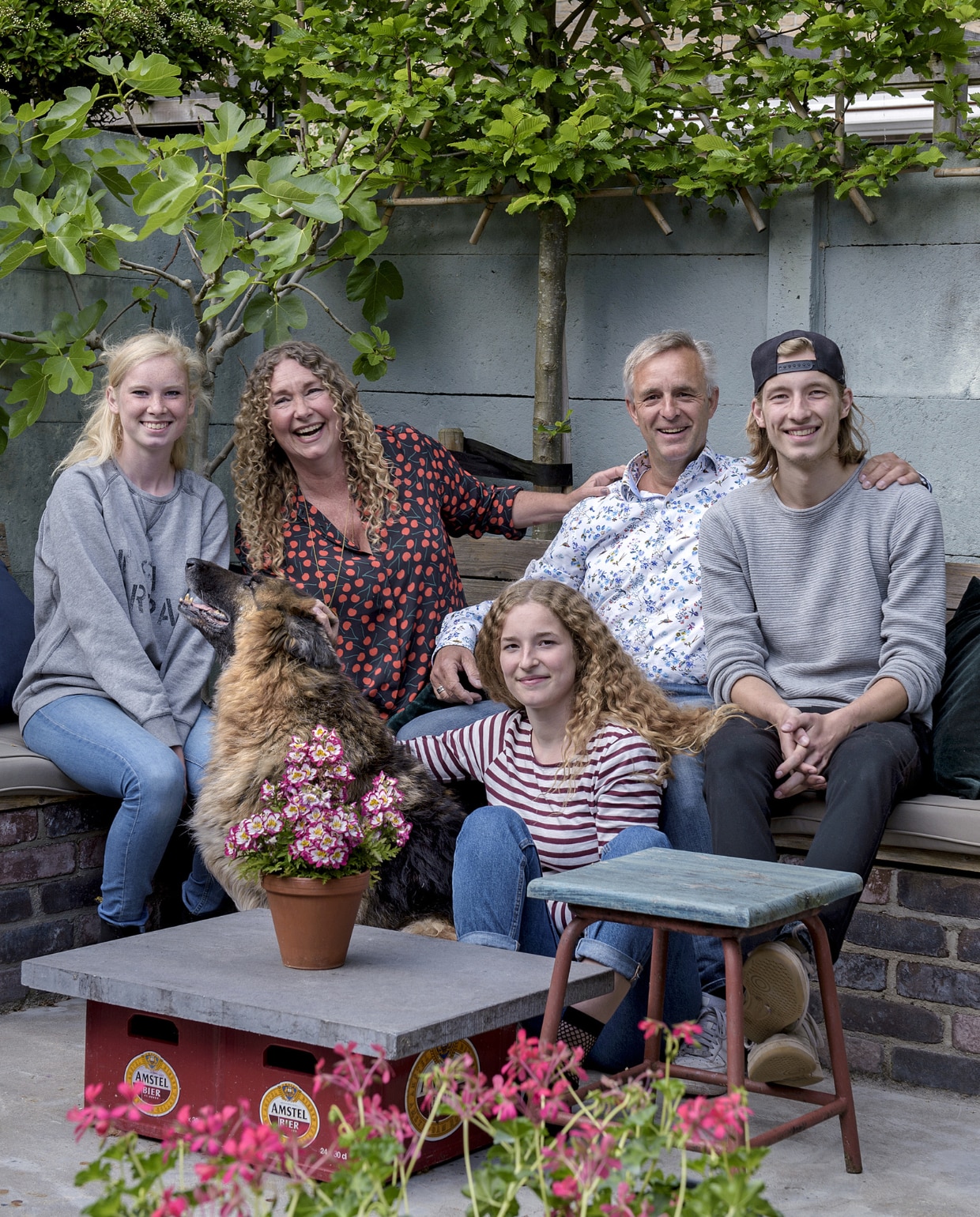 V.l.n.r.: Noa, Wendy, Lola, Koos, Nemo en hond Wolf. In de achtertuin zijn diverse zitplekken ingericht, zoals deze met een gemetselde bank en op maat gemaakte kussens, en een tafeltje van twee lege kratten waarop een deel van het hardstenen aanrechtblad uit het vorige huis. Kruk van de rommelmarkt. vtwonen 01-2019 | fotograaf Ton Bouwer | styling Wendy Romeijn en Monique van der Pauw