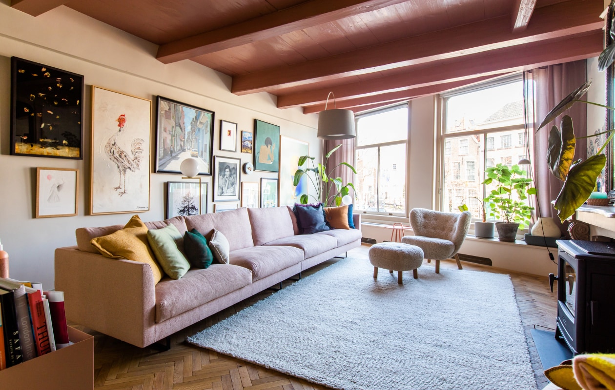Bank en bijzettafel Montis. Kussens Hay, vloerkleed Ferm Living, visgraatvloer Het Leidsch Parkethuis. Verf plafond Blush van Little Greene. Bloempotten, fauteuil en poef &Tradition. Booglamp Foscarini, gordijnen Femkeido, kachel Dover. Kunstwerken (v.l.n.r.): Titus Brein, kip uit Bali, Cuba van Lorenzo de Bruin, zwartwit-foto uit Honfleur, klein rood werk uit Zuid-Afrika, groot werk Isabelle Wenzel. Overig onbekend.