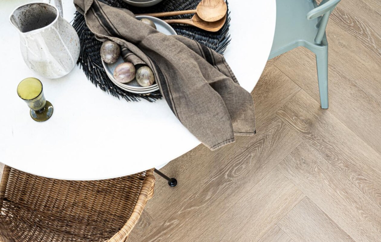 PVC Vloer Herringbone Sun Kissed, 76,5x15,3 cm, dryback per m² € 44,95, click 75x15 cm per m² € 57,95 vtwonen, eettafel en eetkamerstoelen van Woood, grijze borden, kommen, placemats en kruik met handvat Bloomingville, grijze kan wit geverfd Sissy Boy, mand H&amp;M, groen wijnglas, groene waterglazen, houten bestek, groene glazen vaas en theedoek Paviljoen Noord. Overig privébezit. vtwonen 06-2021 | styling Danielle Verheul | fotografie Sjoerd Eickmans