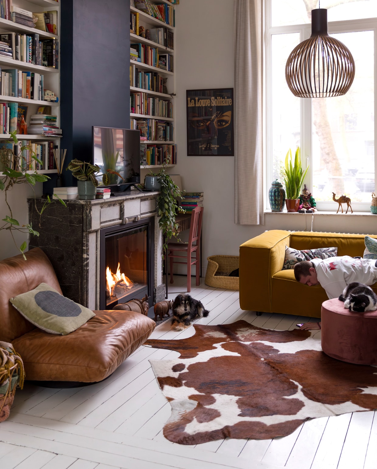 Muurverf schouw Hague Blue van Farrow & Ball, verf overige wanden en vloer Ral 9010. Gordijnen en planken op maat laten maken.  Scheepsfauteuil Woontheater, haard 4M-Fire, potten en planten Van Uytsel. Hanglamp Octo van Secto, potten in vensterbank rommelmarkt, stoel kringloop, vloerkleed koevel van de markt. Bank Dunbar van Fést, poef Sara van Woood, hondenmand Dille & Kamille, poster brocante. Kussens Sissy-Boy en HKliving. Overig onbekend. vtwonen 10-2023 | styling Ilona Jongepier | fotografie Dana van Leeuwen
