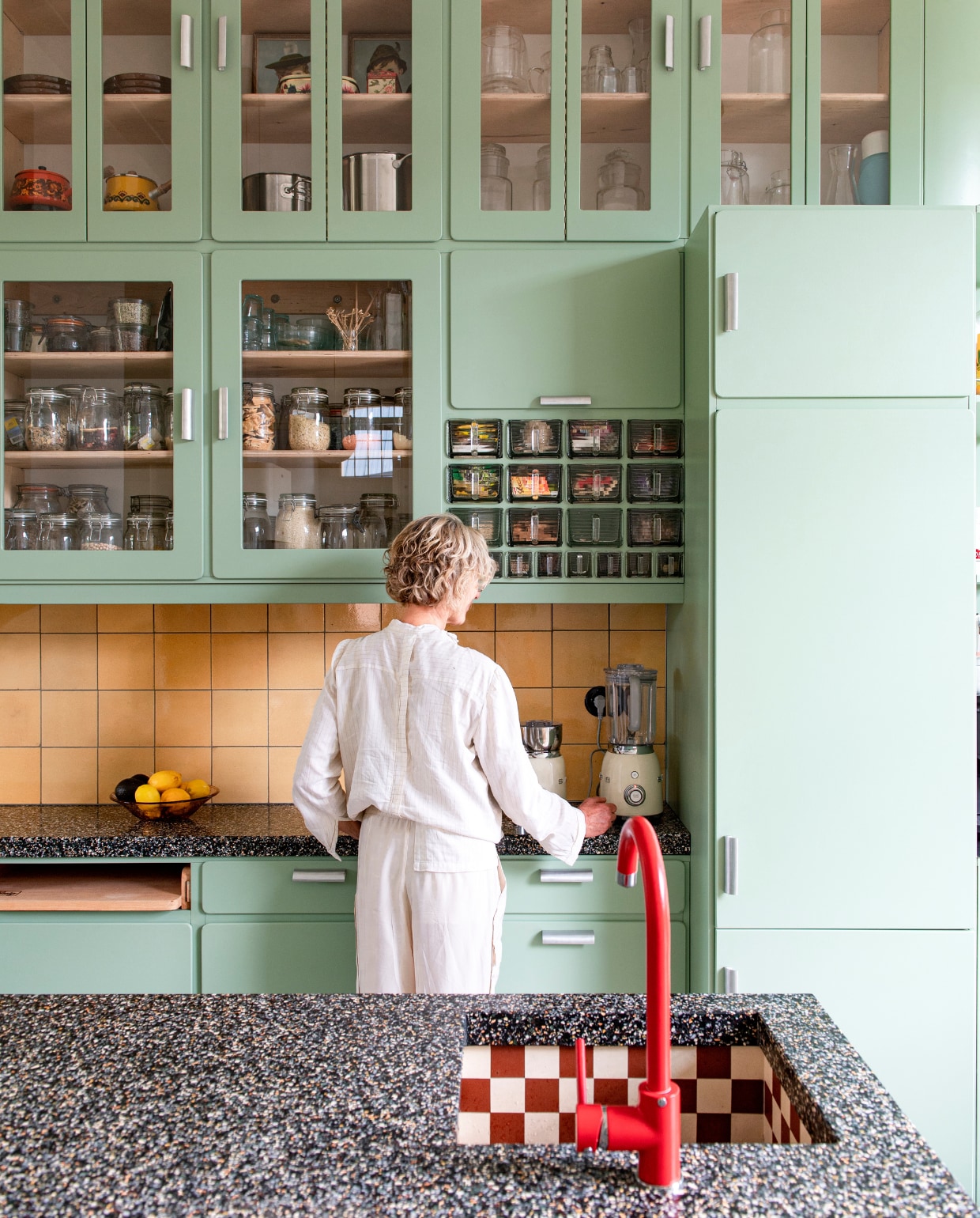 Keuken ontworpen door Studio Daan Hietbrink, verfkleur fronten Room Green 81 van Farrow &amp; Ball. Keukenblad Marcolina Terrazzo en Beton. Rode kraan sanitair.nl. Wandtegels en barkrukken Marktplaats. Vloertegels Winckelmans. Fruitschaal en broodplank uit Frankrijk. Overig onbekend. vtwonen 03-2023 | styling en fotografie Caroline Coehorst i.s.m. Studio Daan Hietbrink