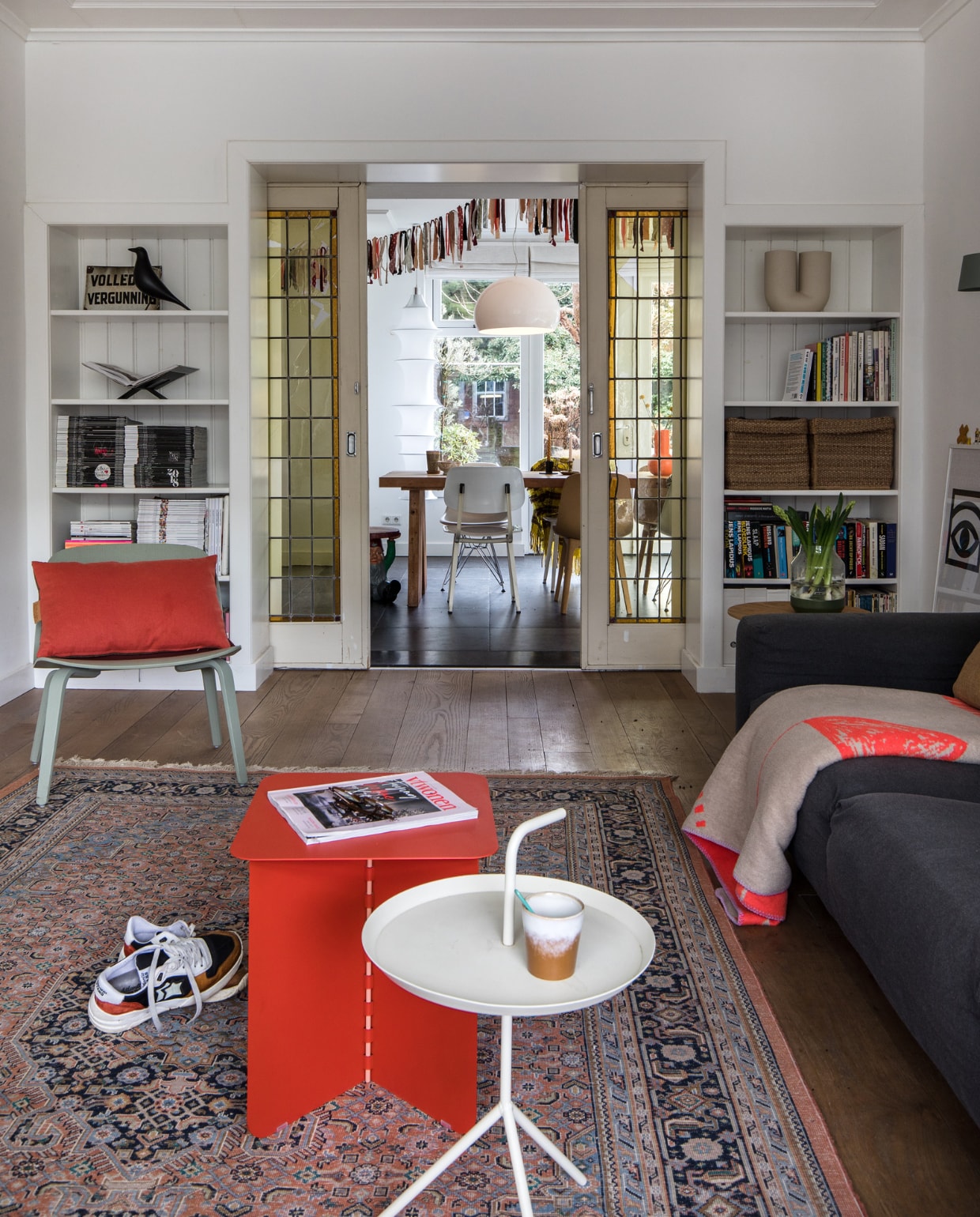 Vloer van Amerikaans eiken lag er al Muurverf wit RAL 9016 Sikkens. Grijze bank gekocht tijdens een garagesale. Plaid op bank Home Sweet Home Adelheid Laden. Vloerkleed kringloop. Sneakers Atlantic Stars. Rode bijzettafel Puik Hinge. Witte bijzettafel Hay. Mok HKliving. Links in kast: drankvergunning uit café van vader, houten kraai Eames House Bird Vitra, boekenstandaard Mendo. Groene Lounge Chair Muuto. Rood kussen Kus-sen. Rechts in kast: vaas Kink Muuto, manden Ikea. Bruine bijzettafel Kwantum. Erop vaas Muuto. Schuifdeuren Marktplaats. vtwonen 10-2021 | productie en fotografie Corine Hoge Bavel