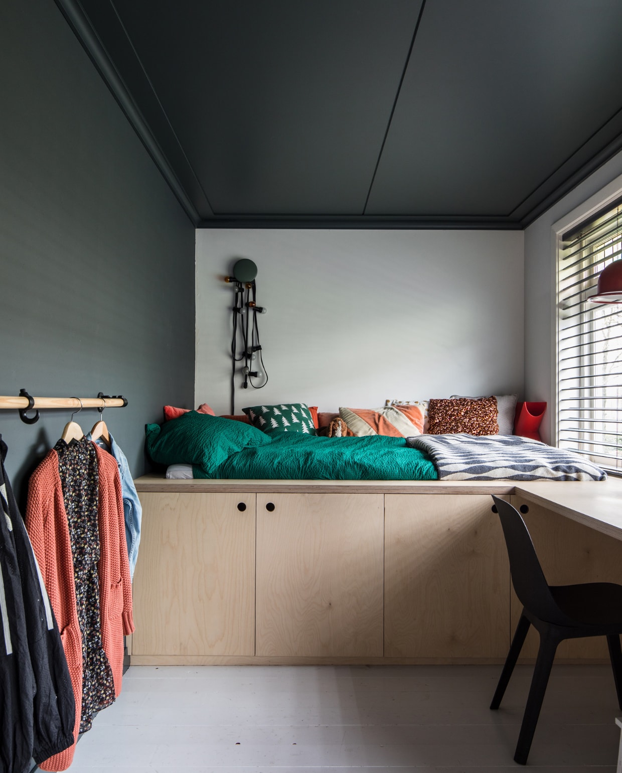 Bed, kasten en bureau zelf ontworpen en gemaakt van multiplex. Kledingrail van oude steigerdelen. Op muur en plafond Full Marble, op de achterwand wit RAL 9016 en op de vloer trappenlak van Flexa. Vlinderjaloezieën Bece raamdecoratie, Hubo. Stoel Odger Ikea. Groene knop Dots Muuto, Flinders. Lichtstreng Kwantum. Dekbedovertrek Hema. Sprei Ikea. Kussen rood, beige, bruin-roze en pluchen beagle Ikea. Groen kussen Ferm Living. Rood kussen La Redoute. Oranje-beige kussen HKliving Rode lamp kringloop, design hanglamp Jieldé brocante markt Lille vtwonen 10-2021 | productie en fotografie Corine Hoge Bavel