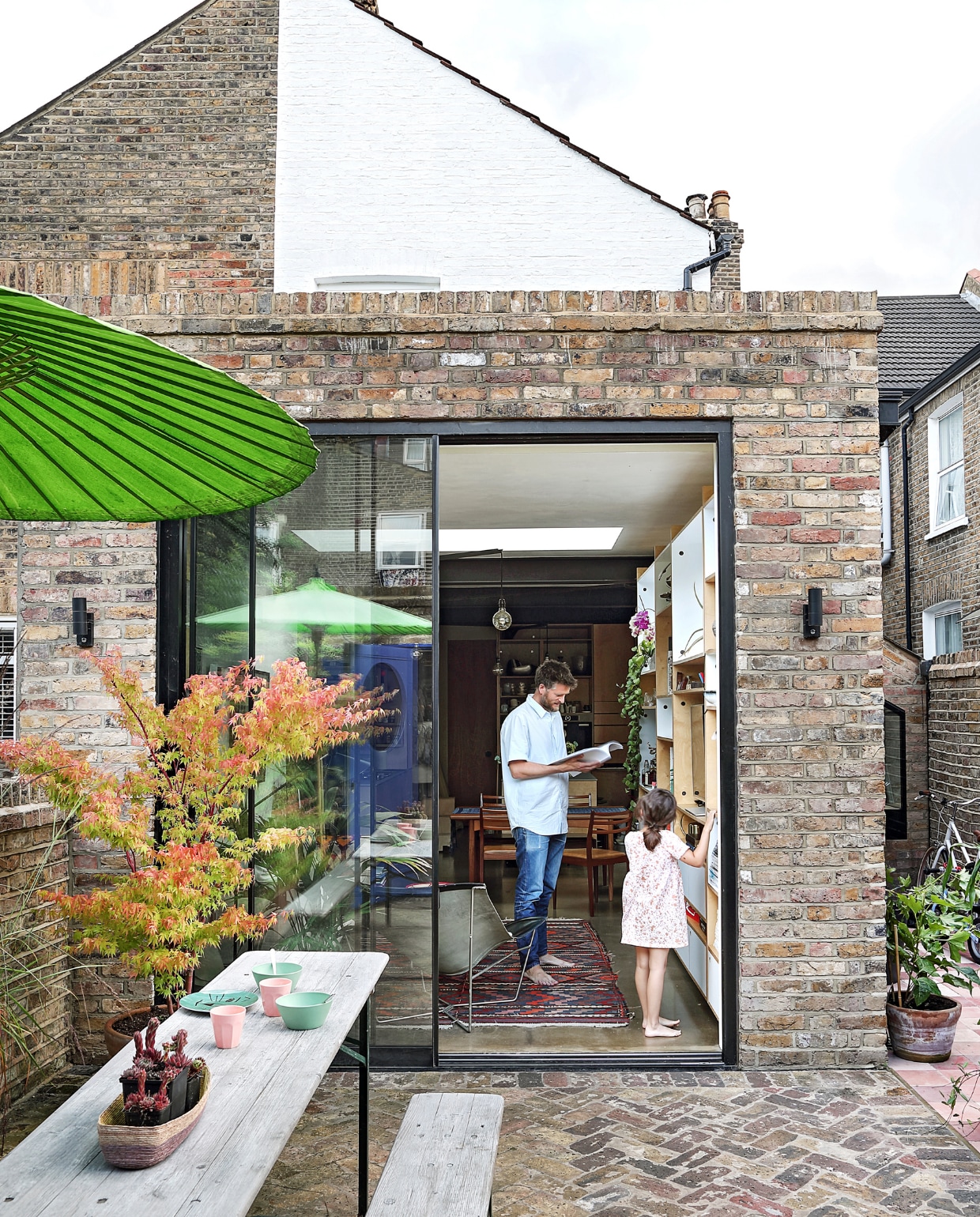 Grote schuifdeuren geven toegang tot de tuin en zorgen voor veel daglicht in dit kleine huis. Sean staat voor een van zijn op maat gemaakte kasten, samen met dochter Freya. Parasol, tafel en bank Scp. vtwonen special binnenkijken 03-2020 | productie Bureaux | styling Someday Studio | fotografie Elsa Young