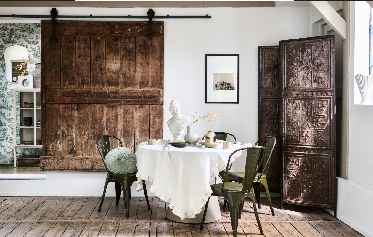 Mdf eettafel Jorre van Woood in Militair Brown, 120x76 cm (Øxh) € 499,-, ijzeren stoel Afternoon van vtwonen € 99,95, porseleinen vaas Tank van BePureHome € 39,95, velvet kussen Halo van BePureHome € 29,95 (basiclabel.nl). Metalen kamerscherm van oude plafonddecoratie, 188x2x194 cm (lxbxh) € 495,- (hetkabinet.nl). Vintage borstbeeld € 98,- (fortuneflea.com). Bekers en borden uit serviesset Picnic in kleur Cashmere van Ferm Living 8-delig € 79,-, metalen borden uit picknickset Yard Picnic van Ferm Living in Cashmere en Olive € 79,- (vtwonen.nl/shop). Linnen tafelkleed met ruche en kunstwerk privébezit. vtwonen 11-2023 | styling Marlies Does | fotografie Sjoerd Eickmans