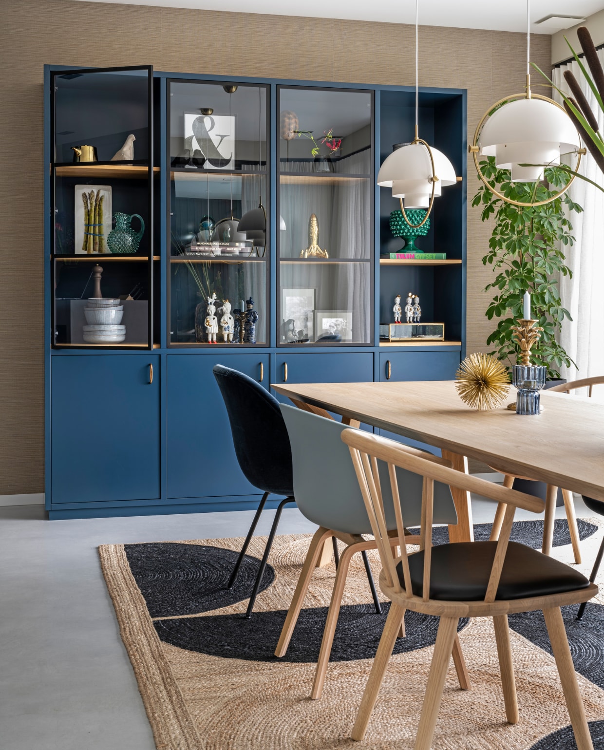 Behang Marsh van Arte, kast gemaakt door Maek Meubels Heerhugowaard geschilderd in Hague Blue van Farrow & Ball, hanglampen Multi-Lite Gubi, tafel Arp Design, vloerkleed Élitis, donkerblauwe stoel en lichtblauwe stoel Hay, houten stoel zwarte zitting Bolia, gouden stekelitem Loods 5, kandelaar palmboom Anna+Nina, waxinelichthouder via Prospectt. In kast: gouden kan HKliving, witte vogel Pigeon van Christien Meindertsma, kunstwerk asperges via Hutspot Amsterdam, blauwe waterkan en groene vaas bollen Mischa Hulshof Alkmaar, grijs servies Broste Copenhagen, print &-teken My Deer Art Shop, witte beeldjes Lammers en Lammers Amsterdam, schildpaddenschild De Museumwinkel, zwart vaasje herkomst onbekend, gouden raket Seletti, fotolijsten  en glazen opbergbox vtwonen collectie. vtwonen 03-2021 | productie CocoFeatures | styling Wendy Verhaegh | fotografie Stan Koolen