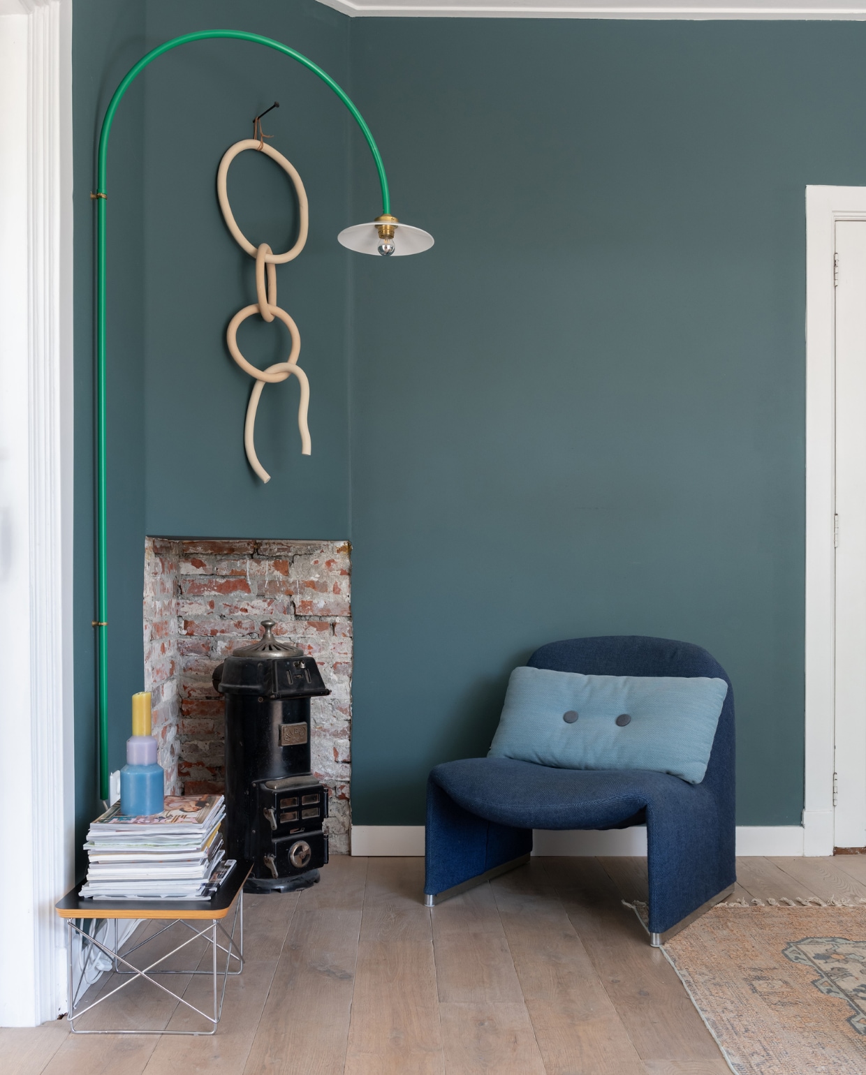 Muurverf Inchyra Blue van Farrow &amp; Ball, vloer Marktplaats, bijzettafel LTR van Vitra, houtkachel tweedehands, wandlamp Muller Van Severen, keramieken wandkunst Lydia Elisabeth Pottery, fauteuil Alky van Artifort, kussen Hay. vtwonen 12-2023 | styling Sharon Roest | fotografie Caroline Martinot