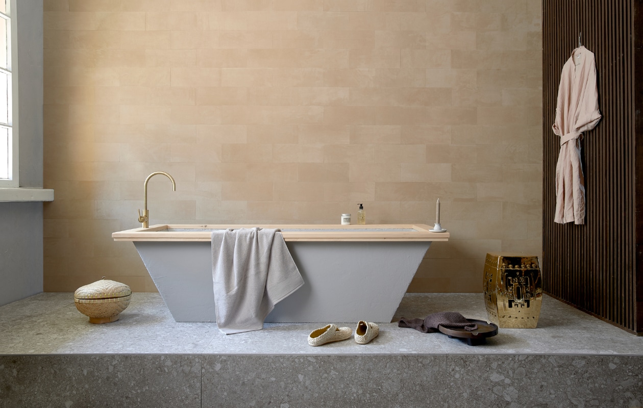 Leren wandtegels uit de Colorado collectie, kleur 3901 desert (ohmannleather.com). Podiumvloer met vloer-en wandtegel Composite Light Grey van vtwonen, 120x120 cm (vtwonentegels.nl). Bad betonlook met houten rand, op maat gemaakt, lattenwand (com-anders.nl). Kraan Grip van vtwonen (vtwonen.nl). Ecru handdoek, schaal, bruine handdoek, linnen ochtendjas (hm.com). Schoenen(sukha.nl). Kruk Temple (polspotten.nl). Rieten mand (loods5.nl). Overig privébezit. vtwonen 10-2020 | styling Fietje Bruijn | fotografie Dennis Brandsma