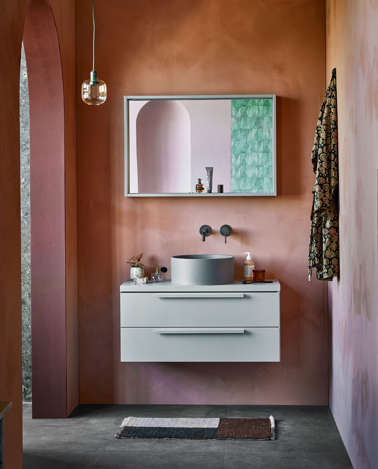 vtwonen collectie (Spiegelkast Handy Beauty in grijs, inclusief ledverlichting en dubbelzijdige spiegeldeuren, 90x16x80 cm (bxdxh) (ook in breedtematen 60 cm en 120 cm en in zand, wit en zwart). Marmeren wastafel Wide in zilvergrijs, 91x20x40 cm (bxdxh) (ook in breedtematen 61 cm en 121 cm, in wit, bruin en zwart en in Top Solid). Kunststof afdekblad in mat grijs, 90 cm. Gelakte onderkast Tidy in mat grijs, 1 lade met handgreep, 90x40x20 cm (bxdxh) (ook in breedtematen 60 cm en 120 cm en in wit, zand, zwart en grijs). Op de vloer vtwonen tegels Raw Antracite, 60x60 cm). vestingh.nl (kleur op de muur Terra Pink No.102, kleur van kolom en rand toog Merlot Mania No. 123), zarahome.com (sloffen), vtwonen.nl (hanglamp Amp van Normann Copenhagen), hay.nl (bruine opbergpot met deksel Container S, vloerkleed Burgundy, tandenborstel Tann, sculpture W&amp;S Moguls Burgundy), nordal.eu (glazen kandelaars, roze vaas, gespikkeld drinkglas, roze marmeren bakje), stories.com (parfum, tubes, sieraden), plieger.nl (wastafelkraan). Overig privébezit. vtwonen 05-2022 | styling Kim van Rossenberg | fotografie Peggy Janssen