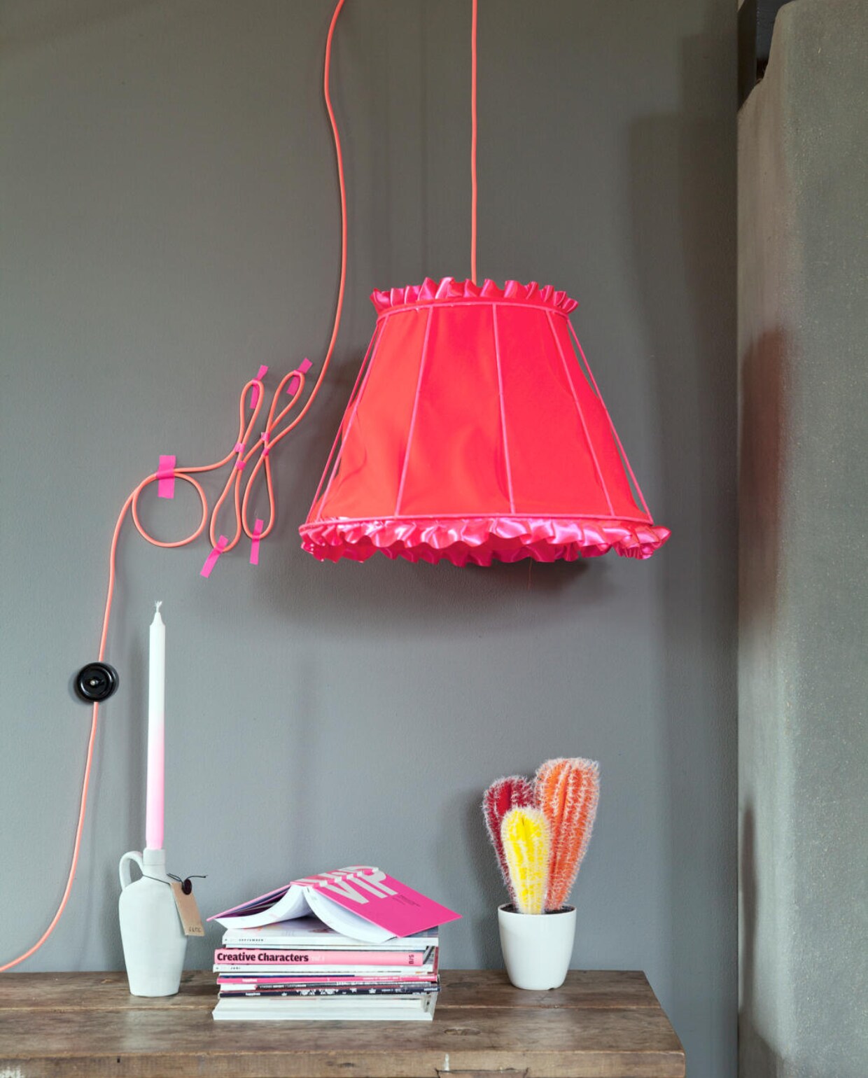 Een oud frame van een lampenkap werd geschilderd in fluorescerende verf Frescolithe kleur Cherry Blossom (Neverlak). Aan de binnenzijde is deze bekleed met stof City Velvet 139 cm breed (Chivasso). Fluo strijkijzersnoer (Snoerboer). Wandschakelaar (Wattnou). Vaasje Cactus (&amp;Klevering). Kaars met fluo dip, vaasje Jack, boek VIP, (Friday Next). Ducktape (Vlieger). Tafel is privébezit. vtwonen 07-2013 | styling Frans Uyterlinde | fotografie Jansje Klazinga