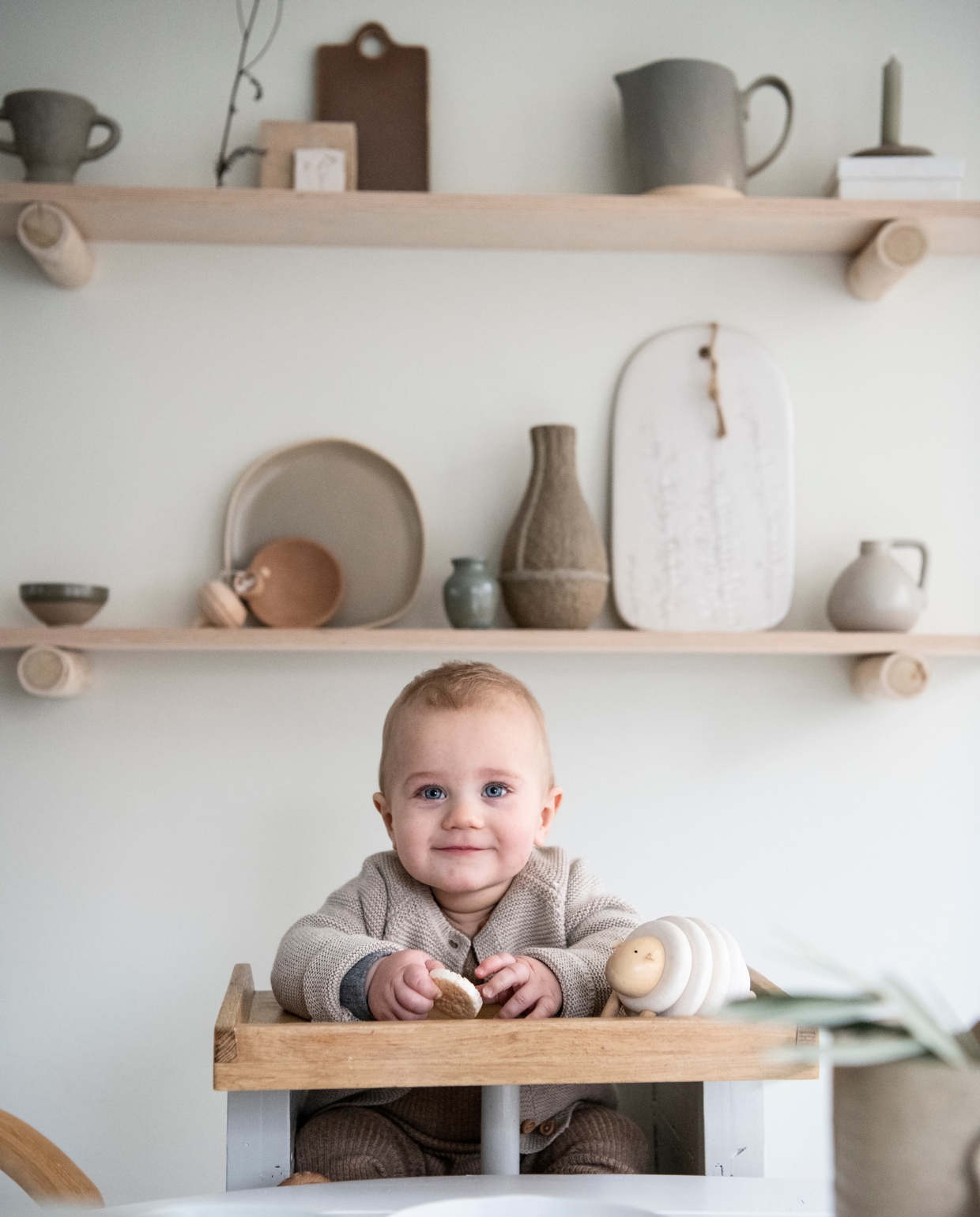 Sol in de kinderstoel van Piet Hein Eek. Houten planken op boomstammetjes van Huis &amp; Grietje. Grijze kom, vaas van papierpulp Serax. Grijs bord Dassie Artisan. Bruine kom Søstrene Grene. Keramieken broodplank Sûr Atelier. Grijze kan vtwonen. Houten schaap Dille &amp; Kamille. vtwonen special DIY 3-2021 | productie Inside Homepage | styling Janneke Peters | fotografie Louis Lemaire