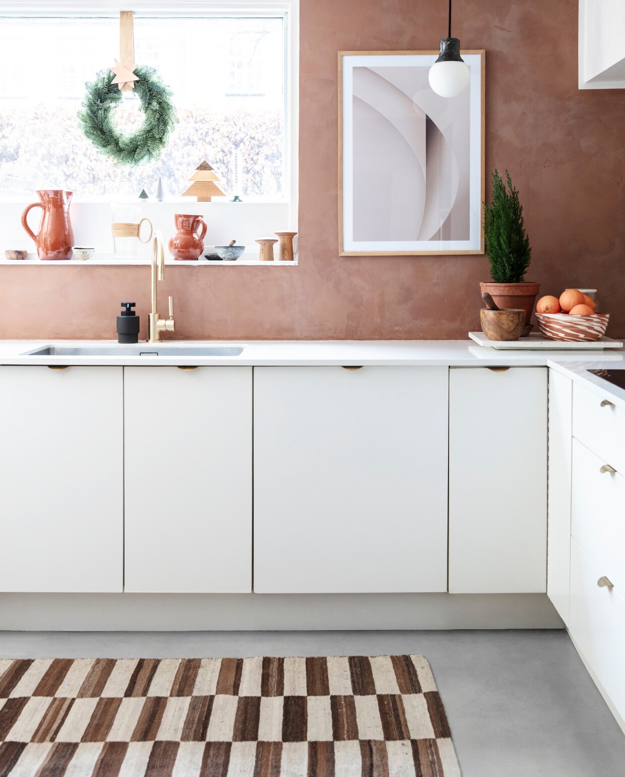 De keukenmuur is geverfd in Lady Minerals kalkverf van Jotun in de kleur Rustic Terracotta. Een groene krans in het raam en een thuja (levensboom) zorgen voor een ingetogen kerstsfeer. Poster van By Garmi. vtwonen 13-2019 | styling Rikke Graff Juel | fotografie Christina Kayser O.