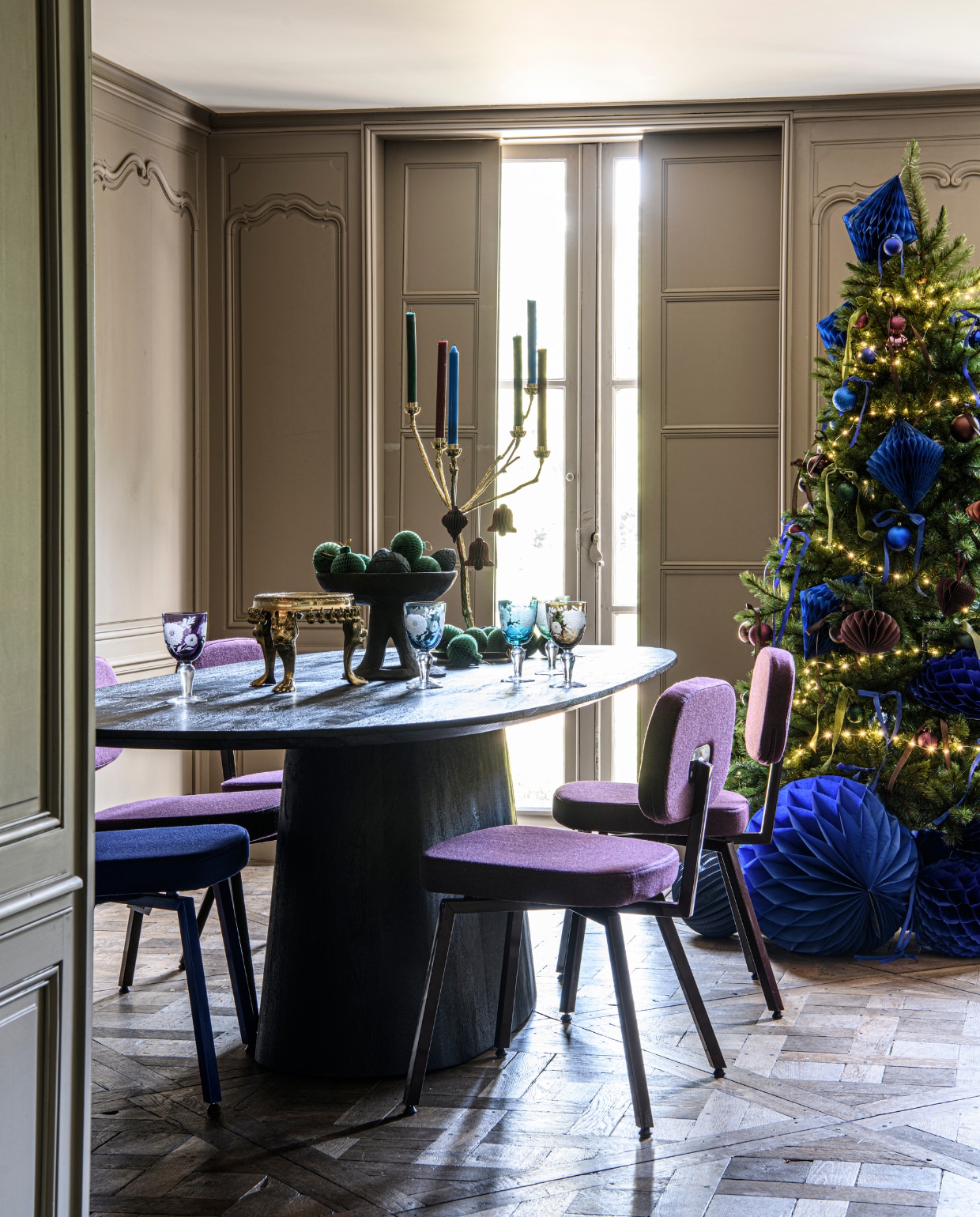 Bruine mangohouten eettafel Aron, 300x110x76 cm  (bxdxh) € 1499,- (eleonora.nl). Eetkamerstoel MK, ultrablauw, met stof Facet Indigo 90 € 269,-, eetkamerstoel MK, zwartrood met stof Purple 78 € 269,-  (demachinekamer.nl). Houten schaal op voet Nagaland € 179,- (rawmaterials.eu). Kleine tafel Royal Lion Altar, gerecycled messing, 33x33x17,5 cm (bxdxh) € 600,- (doing-goods.com). Multicolor wijnglazen Peony, 8,5x17 cm (Øxh), set van 6  € 160,-, vergulde messing kaarsenhouder Twiggy,  50x65 cm (Øxh) € 295,-  (polspotten.com). Dinerkaarsen, diverse kleuren, per stuk € 0,79 (xenos.nl). Kerstballen in ultra- blauw en aubergine, set van 44 € 10,-, bordeaux papieren ornament druppel, set van 2  € 2,50 (hema.nl). Groene honeycomb kerstballen, 8 cm per 4 € 1,95, blauwe papieren honeycomb diamant, 23 cm (h) € 1,55, blauwe honeycomb bal 30 cm (Ø) € 2,20, blauwe honeycomb bal, 50 cm (Ø)  € 4,75, blauwe lampion, 25 cm (h) € 1,80 (partywinkel.nl). vtwonen special kerst 12-2022 | styling Marit Saladini | fotografie Louis Lemaire | assistentie Rosa van Schie, Kiki de Koning