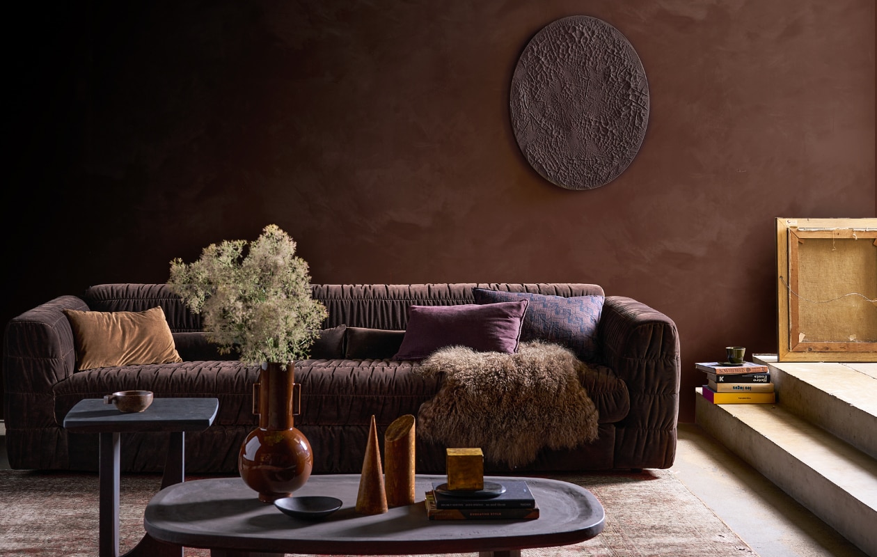 Muurverf kalkverf Aubergine 46 (lauthentique.nl). Fluwelen 4-zitsbank Club Couch in Espresso, 274x108x72 cm (bxdxh) (hkliving.com). Ovale houten salontafel Fille met betonlook finish, 120x75x35 cm (lxbxh), eikenhouten bijzettafel Kurv, gekleurd met gepigmenteerde olie, 45x45x50 cm (bxdxh) (boxworx.nl). Bruine keramieken vaas van HKliving in Espresso, kussen met diagonale strepen van HKliving, 60x40 cm (bxh) (vtwonen.nl/shop). Zwarte metalen schaal, groene beker Grawa (nordal.com). Schapenvacht, fluwelen kussen links, 50x70 cm (iblaursen.nl). Houten geometrische objecten (graublau.nl). Zelfgemaakt canvas muurobject van schilderdoek, 40x50 cm (amazon.nl). Vintage vloerkleed, soortgelijk (rozenkelim.nl). Auberginekleurige fluwelen kussenhoes Astrid van Claudi, 60x40 cm- (wilhelminadesigns.com). Plafondkleur onbekend, overig privébezit. vtwonen 12-2023 | styling Cleo Scheulderman | fotografie Jeroen van der Spek