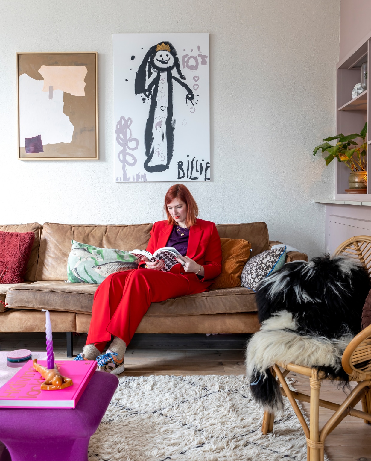 Cathelijne op de bank in haar huurhuis in Wierden. Bank BePureHome. Schilderij links zelfgemaakt, schilderij rechts gemaakt door dochter Billie. Rood en okergeel kussen Loods 5. Vissenkussen Imbarro. Rotan stoel Kwantum. Vacht Intratuin. Fuchsia poef vintage. Roze boek HKliving. Kaarsenhouder zelfgemaakt. Draaikaars Puur Petra. Marokkaans kleed meegenomen uit Marrakech. Kleur kastenwand Light Peachblossom C6B2A7 van Little Greene. vtwonen 13-2023 | fotografie Henny van Belkom