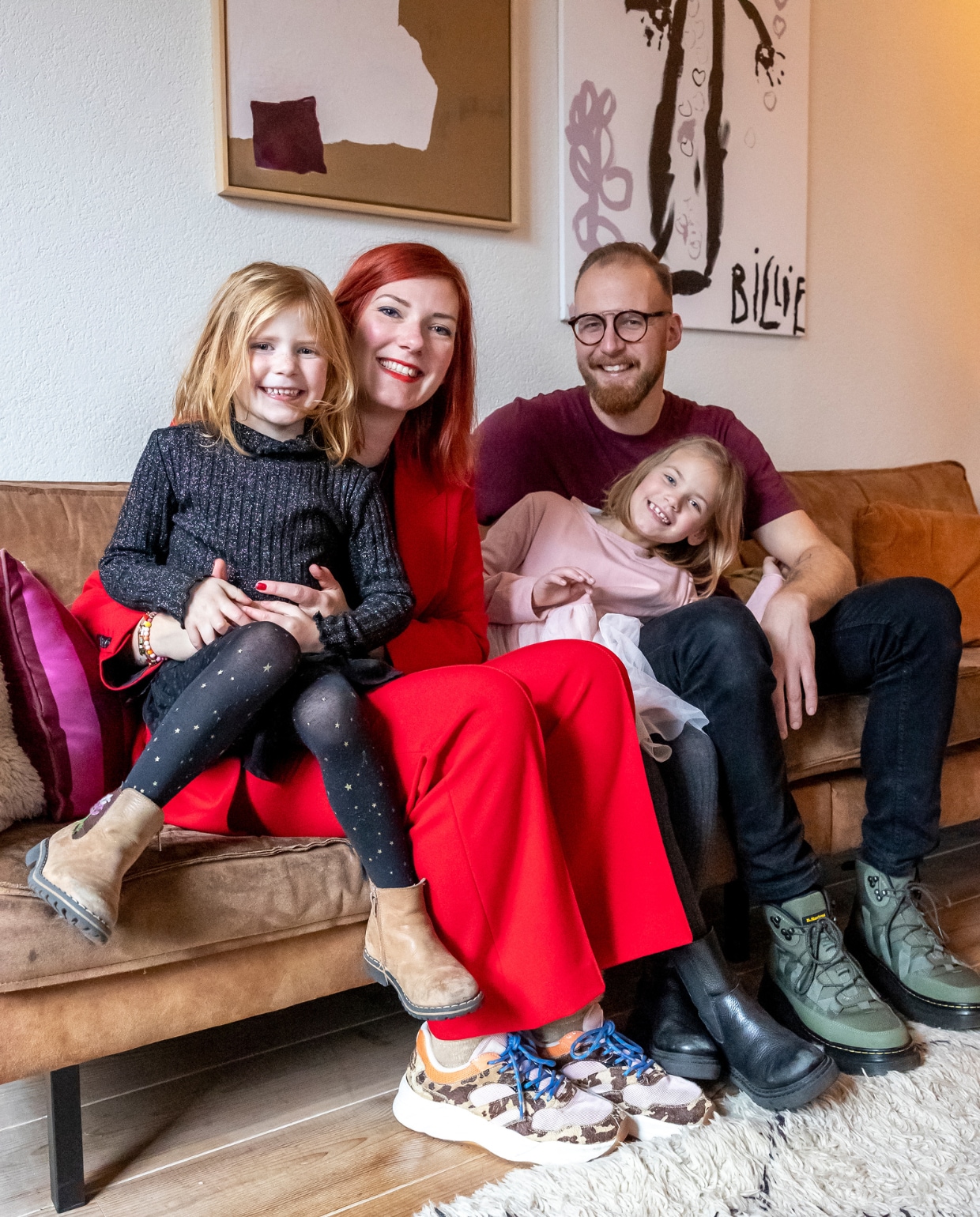 Cathelijne, Maarten, Billie en Fos in hun huurhuis in Wierden. Bank BePureHome. Schilderij links zelfgemaakt, schilderij rechts gemaakt door dochter Billie. Rood en okergeel kussen Loods 5. vtwonen 13-2023 | fotografie Henny van Belkom