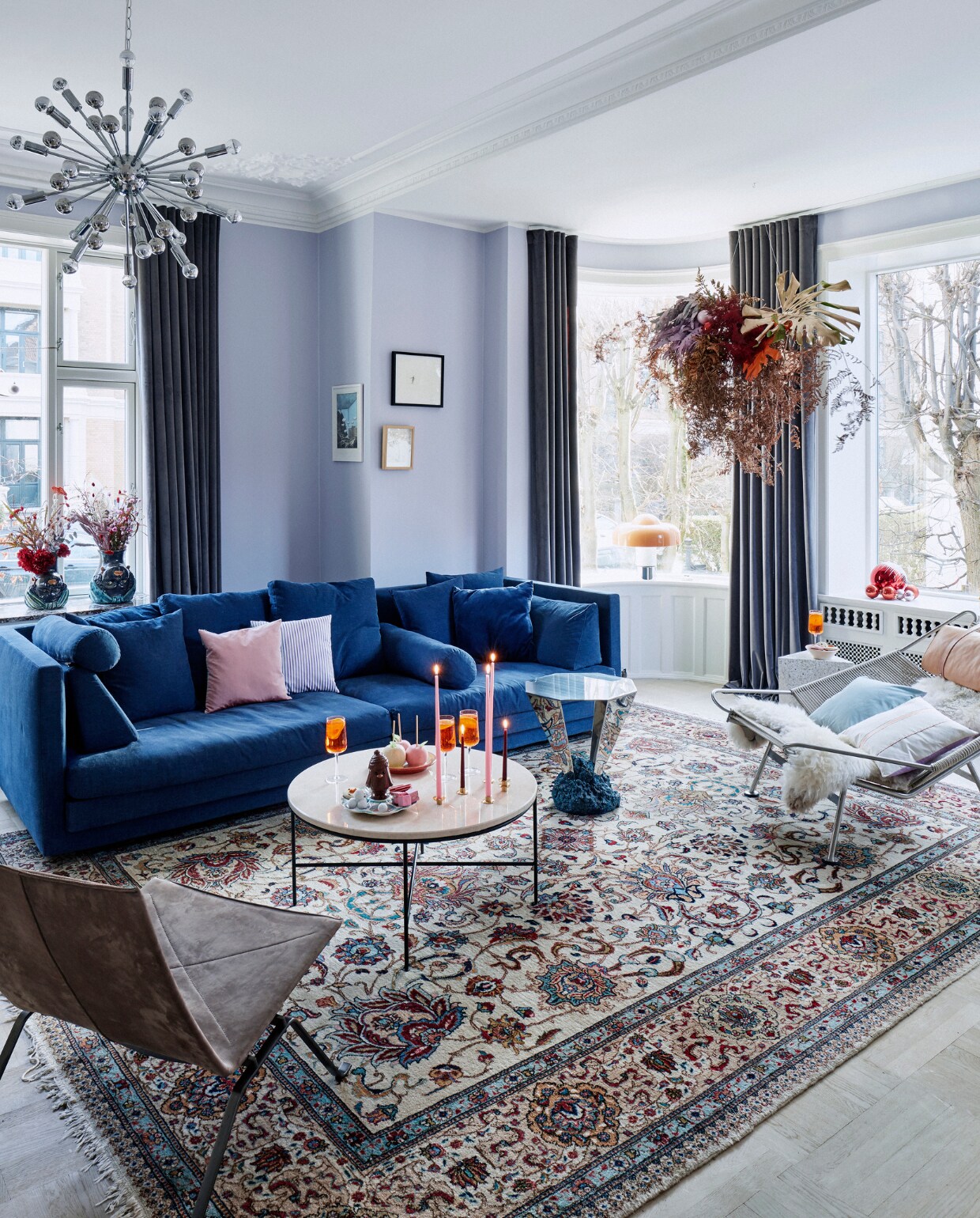 Muurverf s1510-r60b Jotun. Bank Eilersen. Gordijnen in Lavender Haze van &amp;Drape. Donkere fauteuil Poul Kjærholm. Grote salontafel Planner van Fritz Hansen. Hanglamp Sputnik Jolina, vloerkleed erfstuk, hangende decoratie zelfgemaakt, stoel Flagline Hans Wegner. Witte bijzettafel Delta Yume. Muranoglas vazen in vensterbank vintage. Bijzettafel Diamond van Etage Projects. vtwonen kerstspecial 2020 | productie Living Inside | fotografie Martin Sølyst