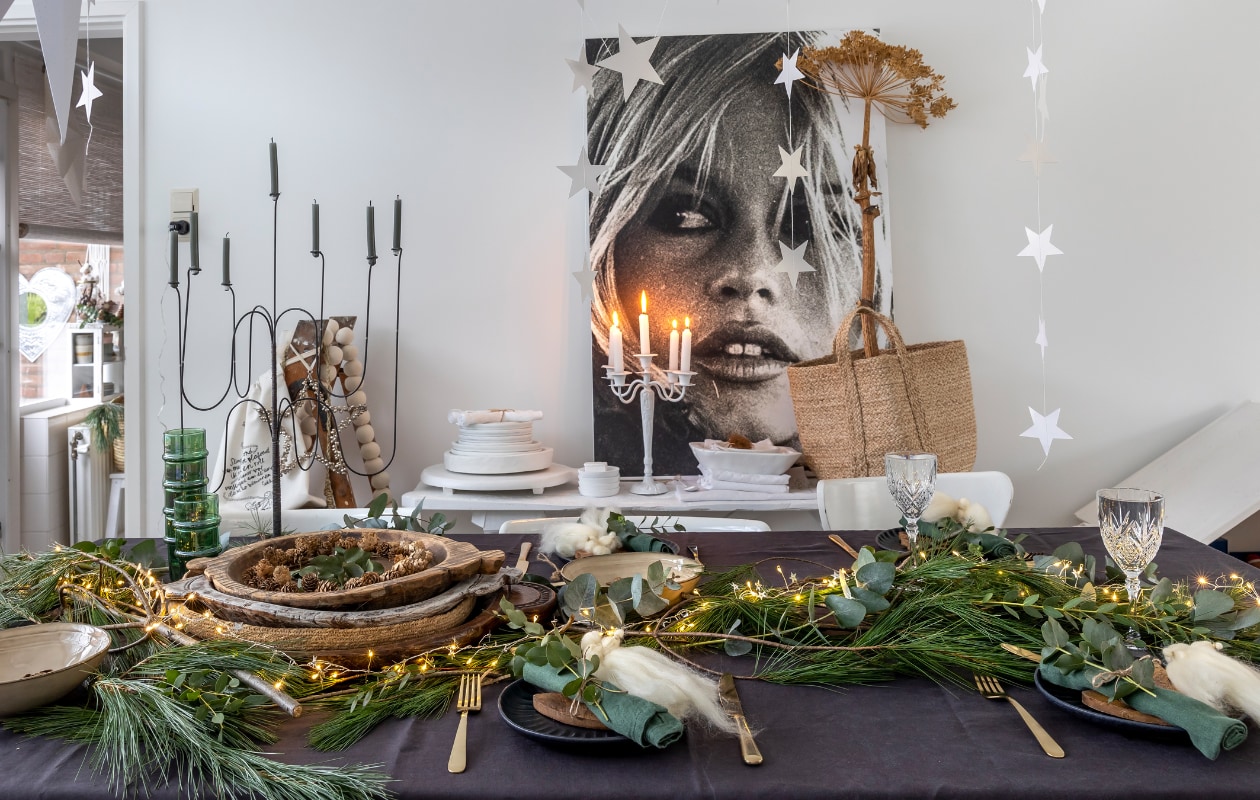 IJzeren zevenarmige kandelaar De Tuinkamer brocante. Mand H&amp;M. Witte ijzeren kandelaar, eetkamerstoelen, houten schilderstrap Marktplaats. Grijs tafelkleed H&amp;M. Gouden bestek Albert Heijn en Sissy-Boy. Witte houten schalen Action (zelf geverfd). Tas Sukha. Groene takken markt. Houten schalen Marktplaats en kringloop. Groene glazen Household Hardware. Groene servetten Søstrene Grene. Zwarte borden Sissy-Boy. Beige schalen Xenos. Wijnglazen kringloop. Sterren Aliexpress. Engelen op borden zelfgemaakt. Overig onbekend. vtwonen 13-2023 | fotografie Henny van Belkom | tekst Ina Babbé