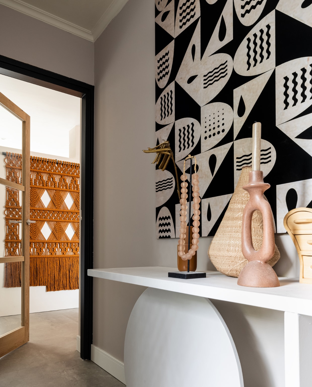 Kleur muur Urban Taupe, Flexa. Deur: privéaankoop. DIY sidetable gemaakt door Richard van Meerveld en Kim Rossenberg in kleur Urban Taupe, Flexa. Kunstwerk ‘Archaïsche kunst’ van Kay Weber, WerkaandeMuur. Staal mat, afmeting 125 x 135 cm. Glass beads ketting uit Ghana, nude O-live. Tonga pot uit Zimbabwe, O-live. Vaasje bruin links Amel glazed ceramic kleur rust, Eijerkamp. Kaarsenstandaard burnt brick, Eijerkamp. Vloer PVC betonlook Dynamic Cemento 609 en plint, Woninginrichting-AanHuis. Overig: privébezit