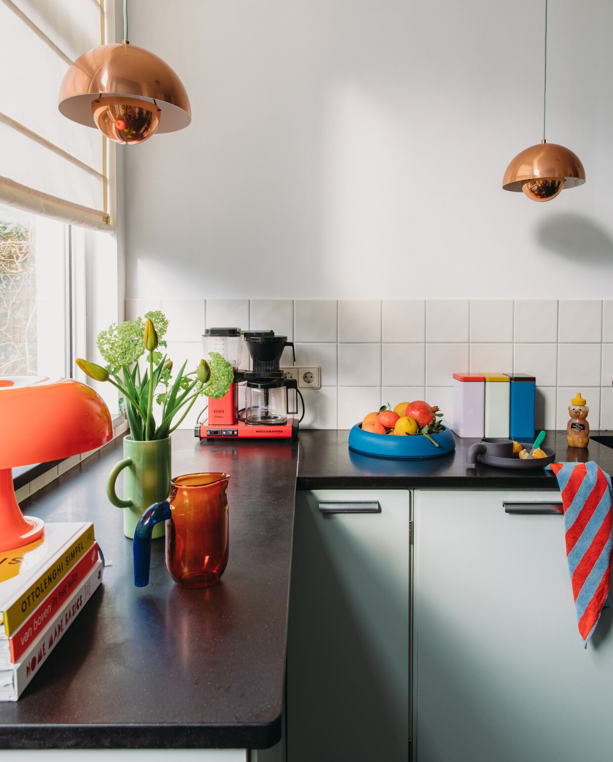 Keuken Piet Zwart van Bruynzeel. Oranje lamp Nessino van Artemide. Kan met bloemen India Mahdavi x H&amp;M Home, waterkan en opbergblikken Hay, fruitschaal Raawii, bord en mok Ineke Hans. Handdoek Marimekko. Hanglampen Flowerpot van Panton. Overig onbekend. vtwonen special binnenkijken 121-2023 | styling StyleStek | fotografie Eva Bloem