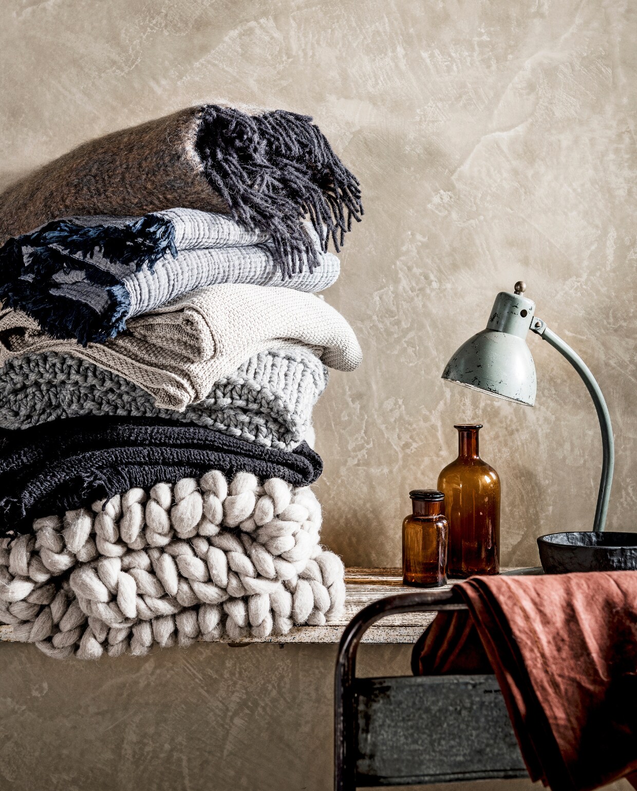 Van boven naar onder: mohair plaid van de Wolwaeren Shore Line collection in kleur Seaweed (Studio Ro Smit). Vergrijsd blauwe gekreukte deken met franje, 150x210 (Hay Amsterdam). Ecrukleurige gehaakte plaid (Dille &amp; Kamille). Grijze gehaakte plaid (H&amp;M Home). Zwarte wafeldeken Svärtan (Ikea). Grof gehaakte wollen deken,100x150 cm (Vacht van Vilt). Metalen stoel, sidetable (Raw Materials). Apothekerspotten (De Weldaad). Bureaulamp (Neef Louis). Zwarte kom Svärtan (Ikea). vtwonen 02-2017 | Styling Danielle Verheul | Fotografie Sjoerd Eickmans