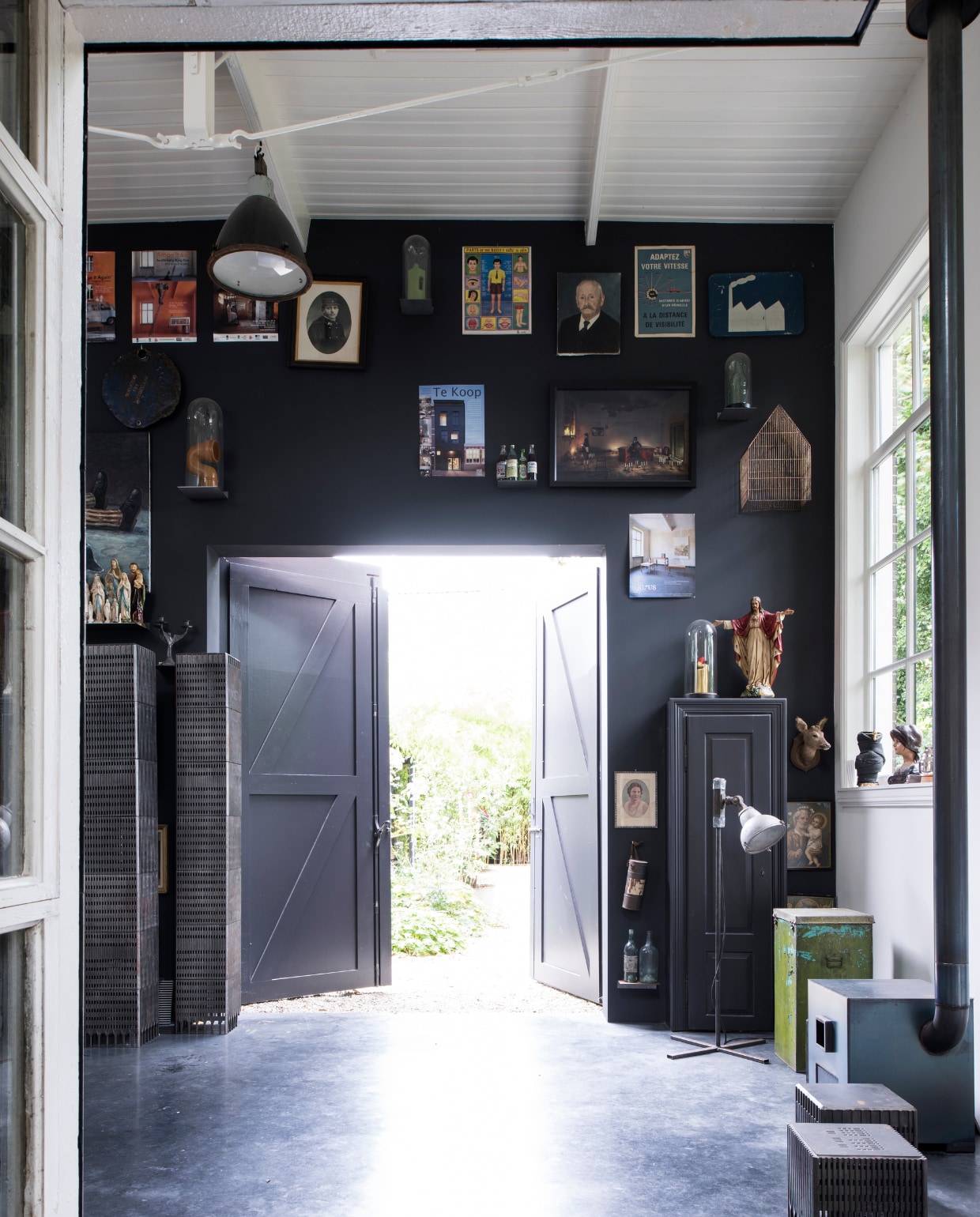 De muur is geschilderd in Rijks zwart grijs van Sikkens. Naast een religieus beeld dat Rolf en Yffi vonden in Frankrijk, staat een gouden wc-eend van kunstenaar Arno Arts. Hij bekleedt flessen schoonmaakmiddel met bladgoud. Industriële lamp gekregen van Niek. Het groene blik komt uit een weeshuis in India waar Rolf met een aantal vrienden twee weken heeft geklust. De houtkachel is geïnspireerd op de voorgevel van hun eerdere huis in Utrecht en gemaakt door Staalstudio. Naast de kachel staat een element van een van Rolfs ontwerpen met de naam WTC light. Verder staat er een vaas van Rolf en een beeld van een Franse brocante. vtwonen 03-2017 | styling Inge van Lieshout | fotografie Louis Lemaire/Insidehomepage