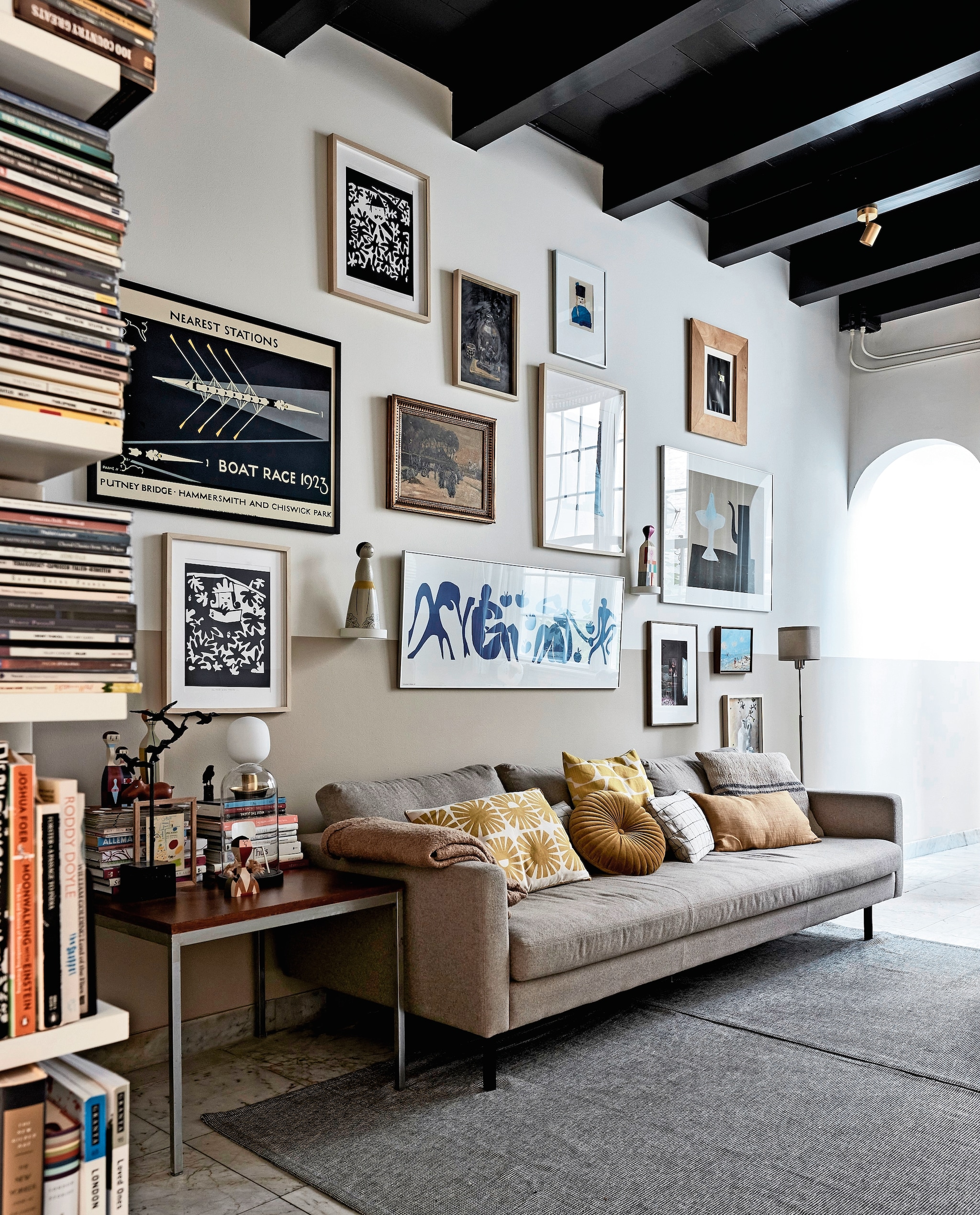 Bank Loods 5. Plaid, rond kussen en effen geel kussen Zara Home. Kussen met zonnetjes, geel-wit kussen, klein kunstwerk op bijzettafel cadeaus. Geblokt kussen zelfgemaakt van Habitat-stof, grijs kussen gemaakt met stof van de markt. Bijzettafel kringloop. Zwart kunstwerk vogels en kleine zwarte vogel familiestukken. Houten beeldjes en leren nijlpaard Vitra. Stolp met model van Revolt-stoel vintage. Tafellamp, vloerkleed en kap stalamp Ikea. Aan de muur: zwart-witte zeefdrukken, schilderij stilleven, persoon met zwarte hoed, groot rechthoekig wit werk in het midden en klein werk vaas met bloemen zelfgemaakt door Heleen. Boat Race 1923 en klein lichtblauw kunstwerk cadeaus. Zeefdruk theepot Klaas Gubbels, ingelijste poster Matisse Ikea, foto Frederique Masselink-van Rijn. Overige makers onbekend. Beeldje gekocht op Bali. Object Vitra. Muurverf  Ral 9010 van Sikkens. Verf lambrisering Pure Mild Sand en plafond Royal Blue van Flexa. Overig onbekend. Beeld vtwonen 02-2024 | fotografie Alexander van Berge