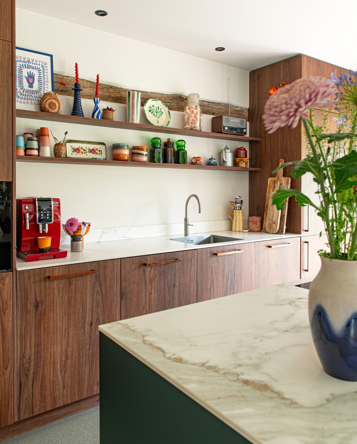 Muurverf Warm White van Pure &amp; Original. Keuken De Haar Keukens, achterwand notenhout kunststof, kookeiland gespoten in Ral 6012, keramieken keukenblad in kleur Estatuario. Rvs kraan Quooker. Koffiezetapparaat De’Longhi, planken Xenos, kan en wijnkoeler Lenta Living, retro radio Denver. Poster H&amp;M, rond vaasje Lovers &amp; Collectors, kaarsenstandaard Loods 5, kaarsenstandaard Que Rico van Via Cannella. Glazen pot erfstuk, 70’s mokken HKliving. Vazen, houten beker, cakeplateau, glazen potten en theepotten @dekringloopzusjes. vtwonen special 131-2024 | productie De Binnenkijkers | styling Jeske Weel | fotografie Caroline Coehorst