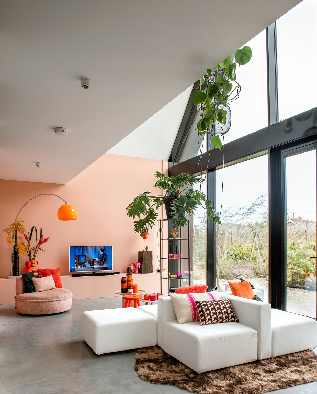 Bank Moduplus, roze bijzettafel Karwei. Kussens en houten blok Nijhof. Stoel Lodge Living, vitrinekast Ikea, vacht Vachten van Terschelling. Roze en oranje kommen en planten De Kaardebol. Oranje hoofd met hoed en oranje vaas links Eigenaar 8. Lamp en  aardewerken vazen vintage. Gietvloer Willem Designvloeren, muurverf Blossom Pink van Le Noir & Blanc. Overig onbekend. vtwonen 10-2023 | fotografie Ton Bouwer