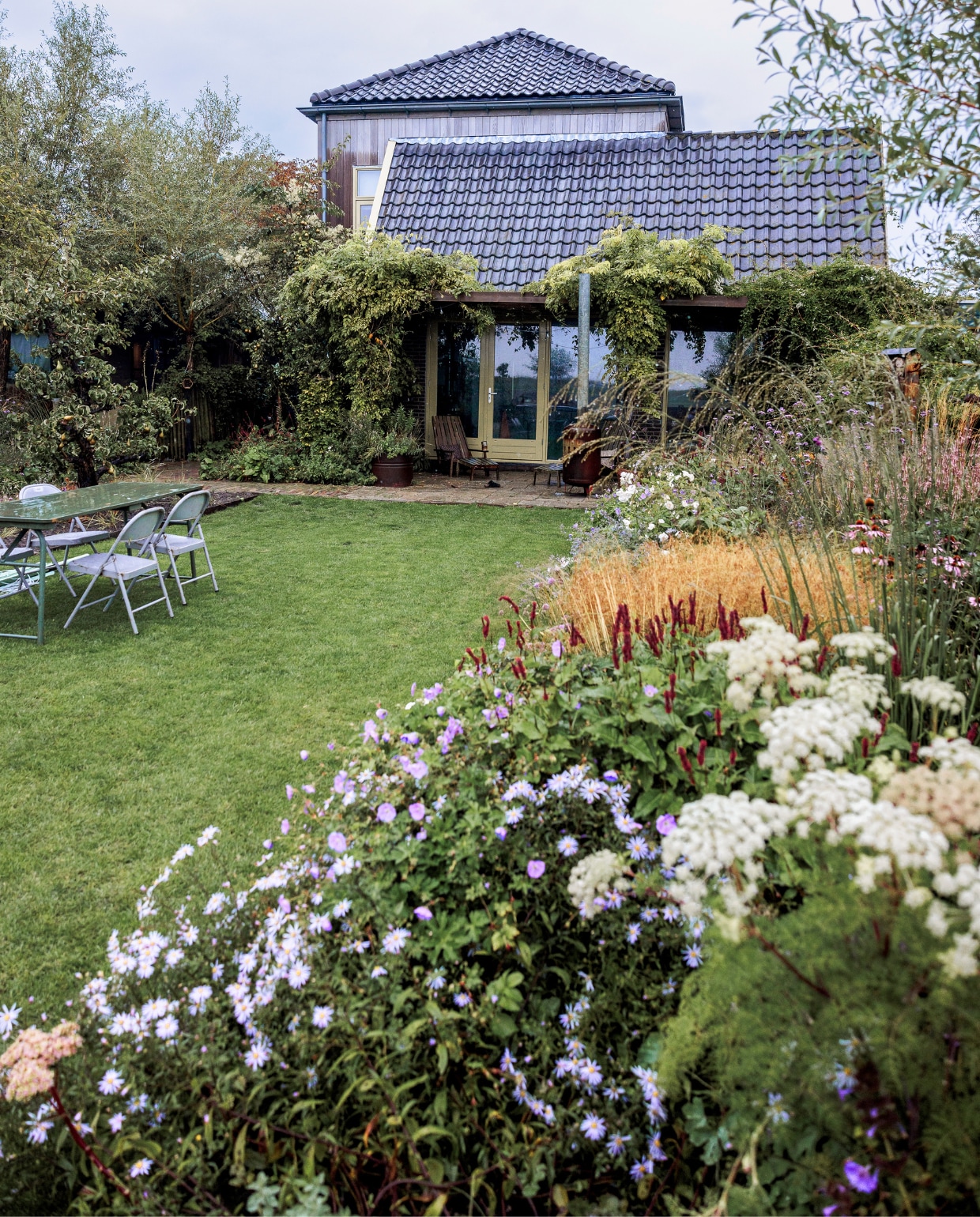 De borders rechts zijn in het voorjaar van 2022 aangeplant met onder meer valeriaan, duizendknoop, ooievaarsbek en aster. Binnen een jaar staat het er al prachtig bij. Op het gras staan een klaptafel en klapstoelen waar Vivian en Mark er meer van hebben, omdat ze makkelijk te verplaatsten zijn naar waar het op dat moment fijn zitten is. Klapstoelen en tafels Combitex. vtwonen tuinspecial 09 | styling Fietje Bruijn | fotografie Ernie Enkelaar