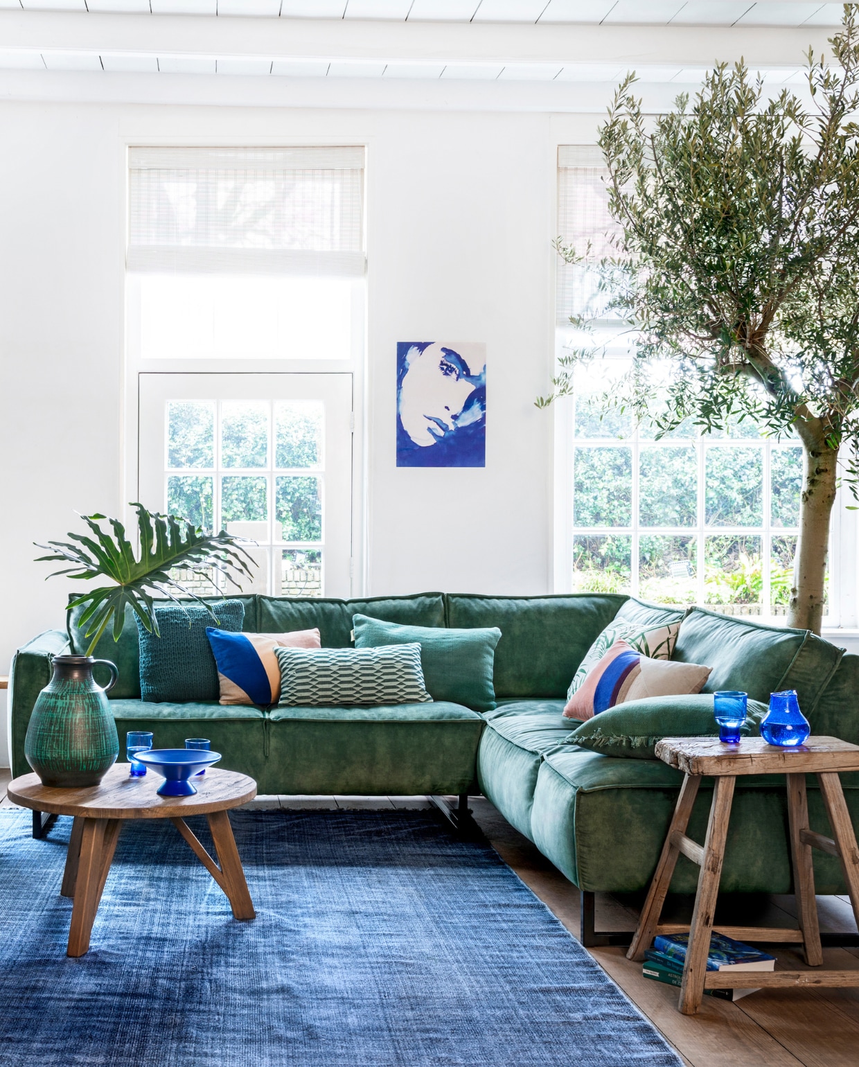 Hoekbank Kick, grote salontafel, kleine salontafel, rotan fauteuil Egg Chair (hetkabinet.nl). Gestreept kussen op fauteuil (sissy-boy.com). Groene linnenmix plaid (hm.com).Vloerkleed Vietnam Days, Stonewashed Blue Cotton Block (aaimadewithlove.nl). Boompjes: olijfboom en Eucalyptus (kwekerij). Naturel-groene mand (manggisliving.nl) met plant Schefflera Elegantissima (tuincentrum). Blauw houten gelakte plantenpot (hetkabinet.nl) met plant Kalanchoë (tuincentrum). Blauwe glazen karaf per 3, blauwe fruitschaal van keramiek, hoge blauwe glazen, lage glazen (hkliving.nl). Grote vintage vaas (mippies.com). Kussens op de bank van links naar rechts; vlakken multicolour (hkliving.nl), langwerpig groen gedessineerd (sissy-boy.com), groen stonewashed kelim (hetkabinet.nl), met bladprint (hm.com), met vlakken Patch (hkliving.nl), groen stonewashed Kelim (hetkabinet.nl). Poster (hkliving.nl). Bankje, grote plantenpot links, houten kruk en boeken privébezit. vtwonen 05-2019 | styling Moniek Visser | fotografie Stan Koolen