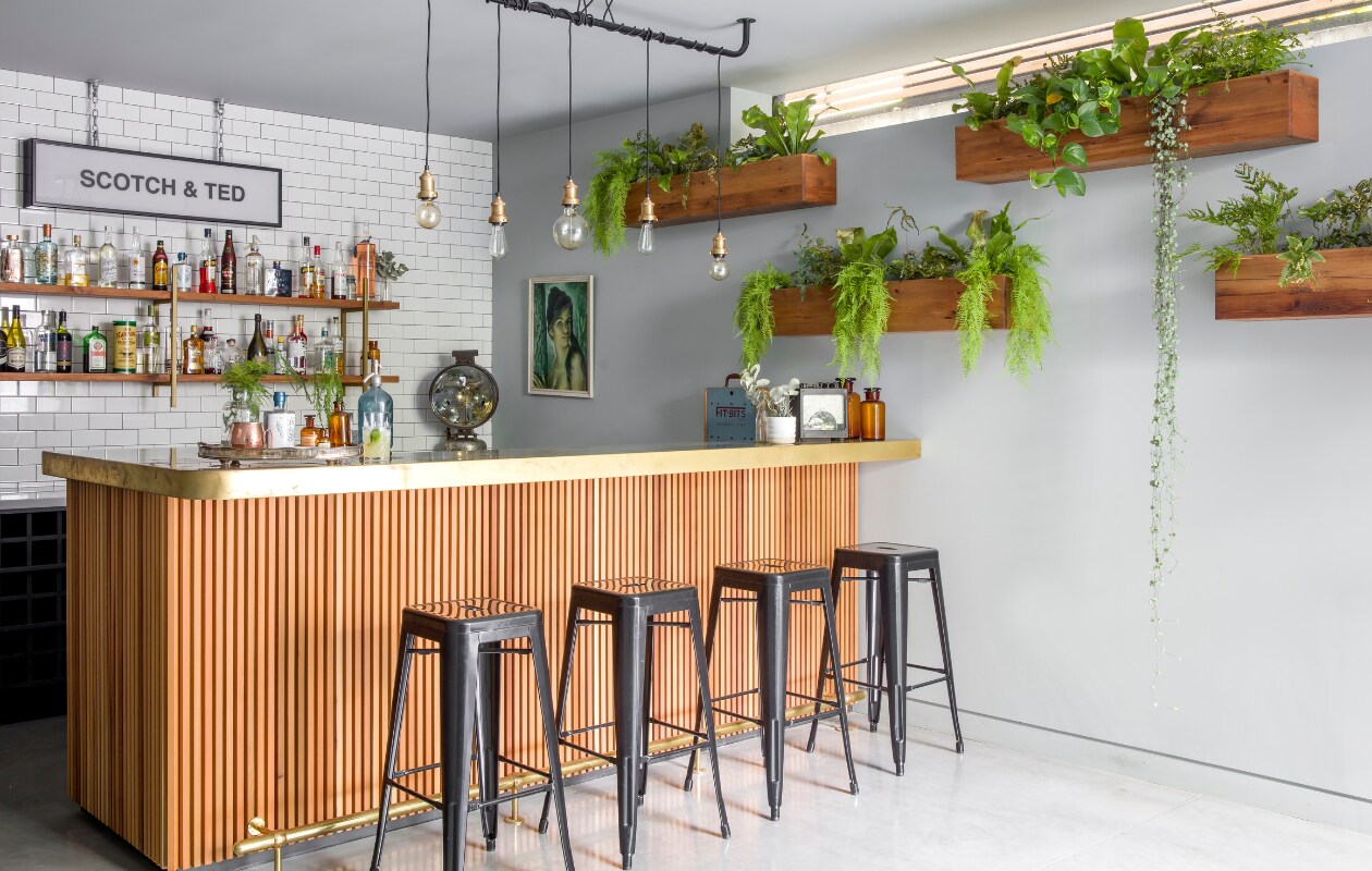 De cocktailbar in de kelder. Het bord aan de muur verwijst naar Sommers grootmoeder: ‘Ze had twee grote liefdes, mijn grootvader Ted en Scotch’. De bar is op maat gemaakt door Jason &amp; Co. Aan de muur houten plantenbakken met veel hangende planten die de ruimte een natuurlijke uitstraling geven. vtwonen 01-2020 | fotografie Rei moon/living Inside