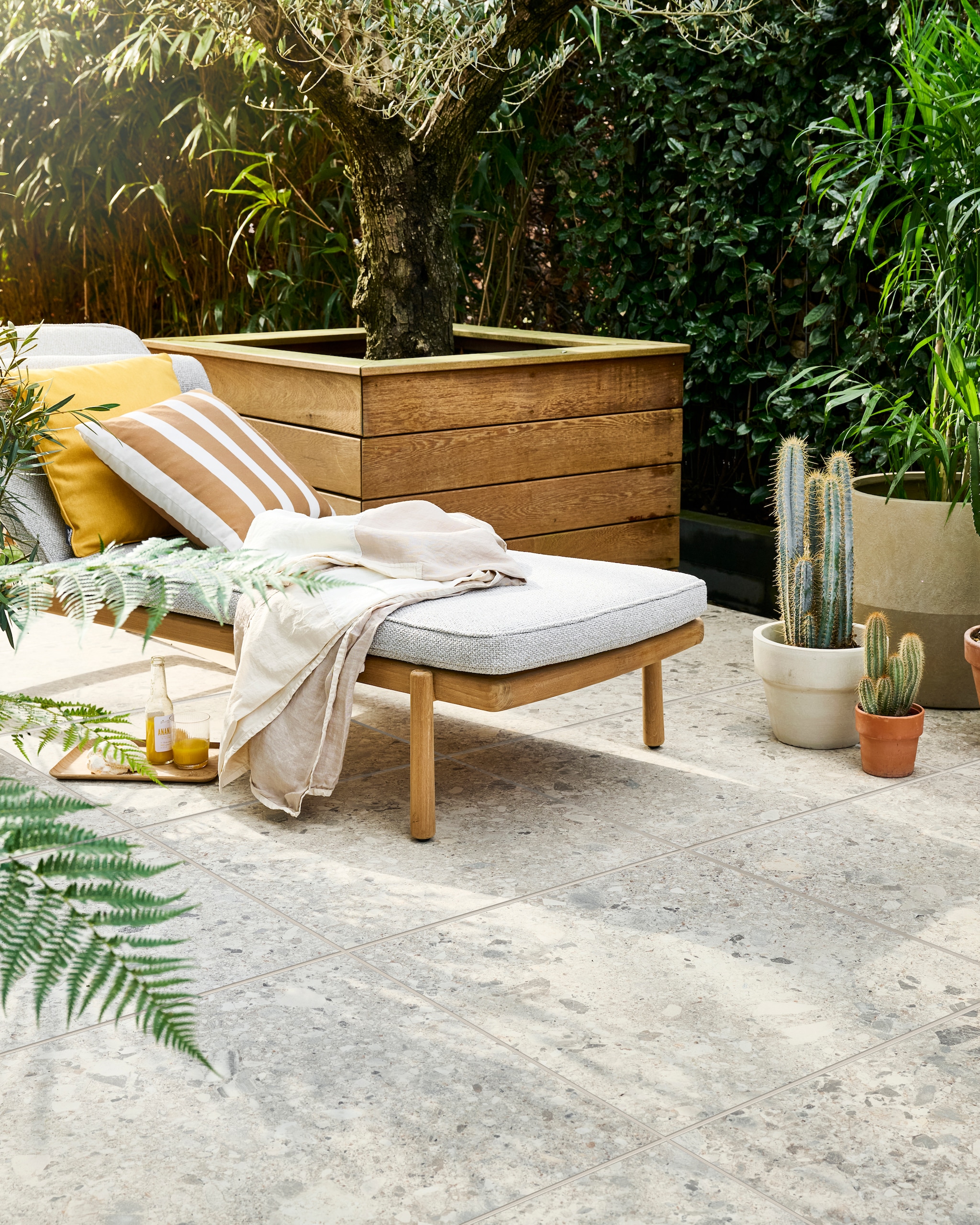 Tuintegel Solostone Uni Composite Mix Beige, 70x70 cm, per m² € 74,50, ook verkrijgbaar in Mix Grey. M.m.v. dacks.nl (ligbed met matraskussen Pure Sofa van Tribù), intratuin.nl (geel kussen, gestreept kussen, terracotta bloempot), vtwonen.nl/shop (plaid, witte bloempot van Serax), zarahome.com (dienblad, glas). Overig privébezit. vtwonen 05-2024 | styling Marlies Does | fotografie Sjoerd Eickmans
