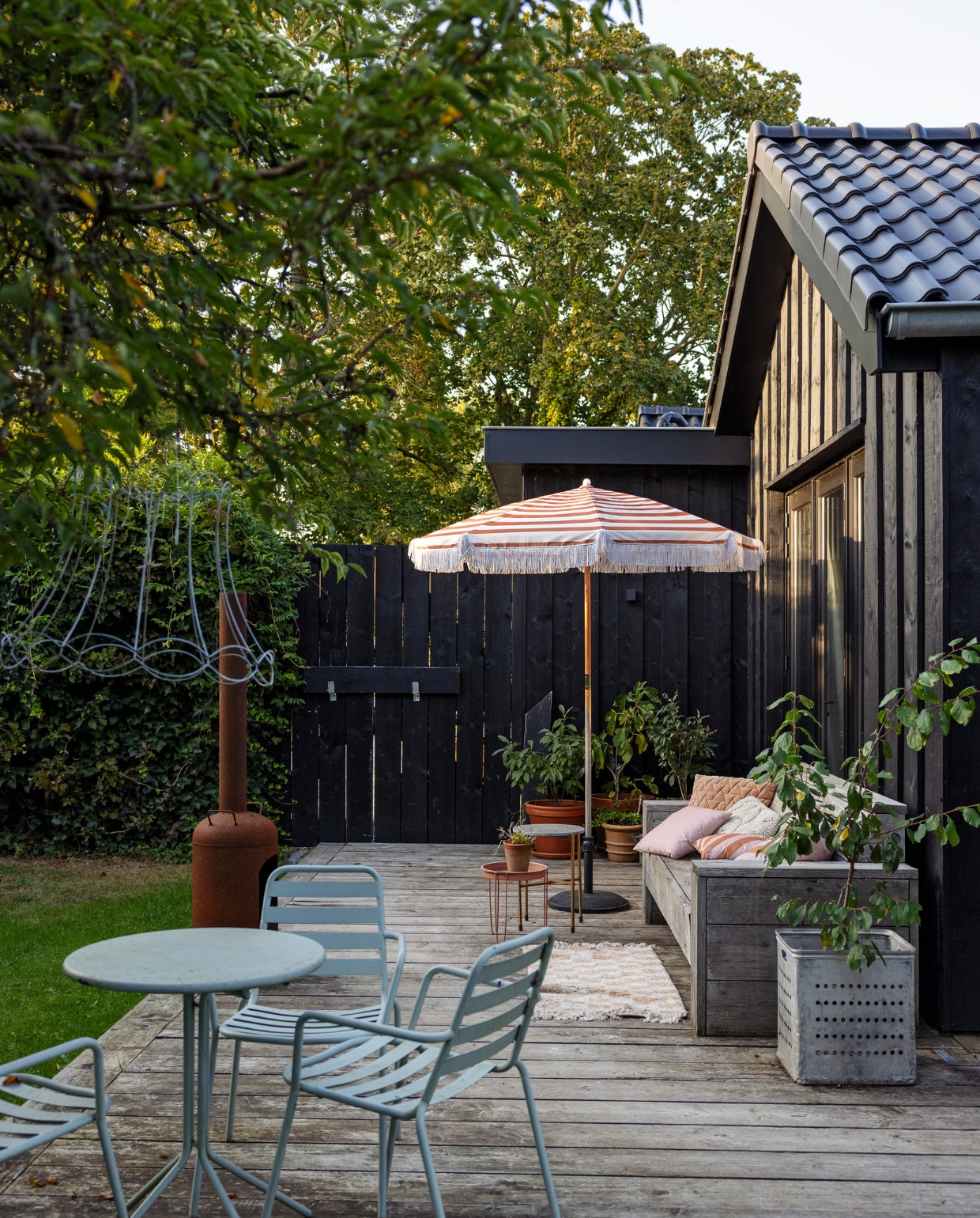 In de potten staan planten die elders uit de tuin werden gehaald.Zinken bakken van brocantemarkt in Frankrijk, met in de voorste bak een perzik. Bank door timmerman gemaakt van steigerhout, houtkachel Marktplaats, in de boom een frame van een oude lampenkap. Tuinset, parasol, tafels en kussens Kwantum. vtwonen Tuin 10 | styling Ilona Jongepier | fotografie Dana van Leeuwen