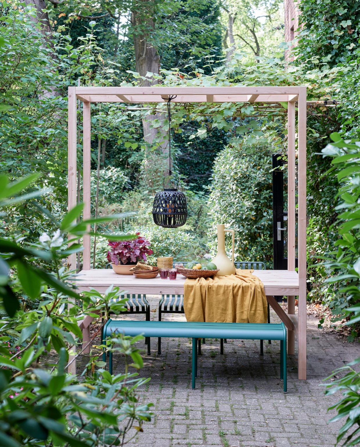 Eettafel met pergolaa. Totaalbeeld eettafel met pergola. Groene Fermob Luxembourg tuinbank € 485,-, groene stoelen Hay pallisade chair groen € 269,- p/st (vtwonen.nl), tafelkleed okergeel gemaakt van okergele stof € 8,95 p/m (patickkarteris.nl), hoge okergele vaas € 35,- (kwantum.nl), zwarte bamboe hanglantaarn € 20,- (intratuin.nl), sisal schaal € 19,99 (hm.com), okergele diepe borden Glaze € 4,- p/st (kwantum.nl), okergele schaaltjes € 4,99 p/st, okergele schaal € 5,49 (intratuin.nl) vtwonen Tuin 05 | productie Corrien Flohil | styling Moniek Visser | uitvoering Johan Snoodijk | fotografie Sven Benjamins
