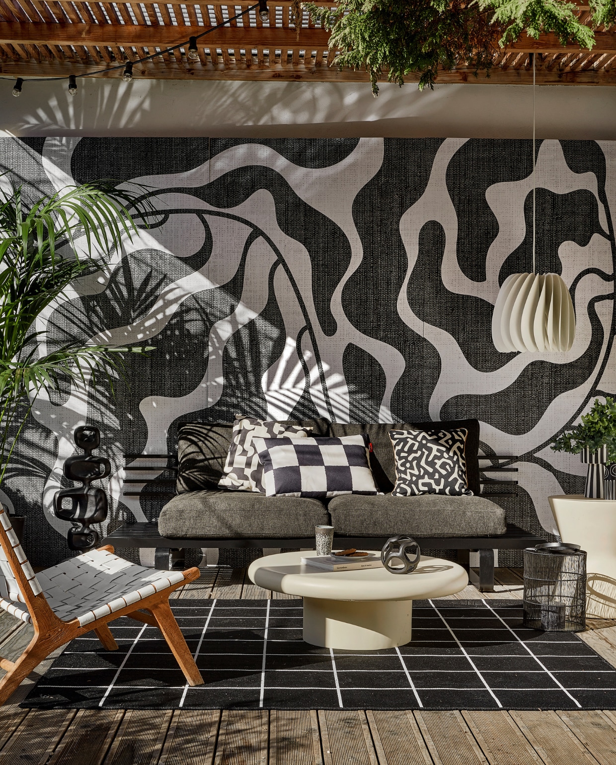 Behang River black &amp; white van Wrap the Wall, per m2 € 150,-, minimale afname 4 m2 (behangfabriek.nl). Zwarte aluminium buitenbank € 1339,80, antraciet tuinkussens, per set € 675,75, zwarte aluminium sculptuur € 295,-, mok 70s, per 2 € 19,99 (hkliving.com). Buitenvloerkleed Svallerup, 200x200 cm € 79,95, hanglamp Skymningen € 79,95 (ikea.nl). Ivoorkleurige polyester bijzettafel, 85x30 cm (Øxh) € 599,-, bijzettafel Pot-Table in Ivory-Slim € 395,- (householdhardware.nl). Zwart-wit kussen Cutro € 10,- (kwantum.nl). Zwart-wit geblokt kussen King Pillow € 55,- (fatboy.com). Kussen Nara € 59,90 (ateliersem.nl). Zwart-wit gestreepte pot van Oyoy € 69,95, aardewerken vaas Troy van Bloomingville € 12,95 (livvlifestyle.nl). Zwart aluminium bolvormig ornament € 31,95, lantaarn € 96,95 (ptmd.nl). Fauteuil met gevlochten zitting privébezit, soortgelijke bij Kave Home (kavehome.com). vtwonen 06-2023 | styling Kim Timmerman | fotografie Peggy Janssen