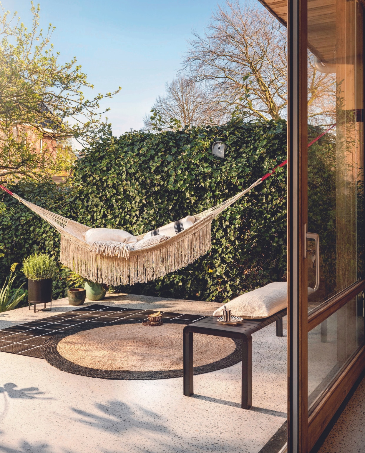 Hangmat Path in Light Sand, gerecycled plastic, 320x140 cm (lxb) € 229,-, zwarte stalen bloempot Bau L, 28x38 cm (Øxh) € 79,- en beige kussens Nettle van Ferm Living, brandnetel en biologisch katoen € 139,-, metalen tuinbank Vondel van Zuiver, 129x45x45 cm (bxdxh) € 599,-(vtwonen.nl/shop). Vezelcement vogelhuis Birdy van Eternit € 110,-(extravert.nl).Gebreide wollen kussens Frills € 49,- (zuiver.com). Geruit buitenkleed Svallerup, 200x200 cm € 69,95 (ikea.nl). Juten vloerkleed Rondo, 180 cm (Ø) € 85,- (hetkabinet.nl). Gestreept wollen kussen € 95,-, keramieken mok € 12,95, zwart-wit keramieken bord € 17,50, keramieken kom € 16,50 (householdhardware.nl). Overig privébezit. vtwonen 07-2024 | styling Marianne Luning en Kelly van Gendt | fotografie Stan Koolen