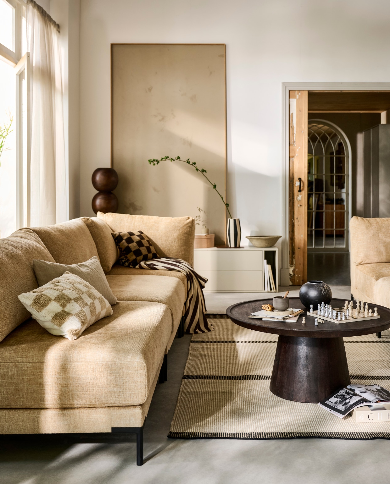 Elementenbank Couple in stof Butter Melange, zoals hier samengesteld: hoekelement links € 949,-, loveseat € 1049,- en hoekelement rechts € 949,-, verkrijgbaar in verschillende stoffen. Bijzettafel Coffee A Gogo, mangohout en walnootfineer, 90x40 cm (Øxh) € 499,-. Loveseat Couple in stof Butter Melange, 100x100x89 cm (lxbxh) € 1049,-, verkrijgbaar in verschillende stoffen. Grenenhouten tv-meubel Settle Down in Dust, 200x44x45 cm (bxdxh) € 499,-.M.m.v. vtwonen.nl/shop (vloerkleed van Linie Design), velv.nl (kussens Eve en Pure, mok, minivaas op tv-meubel), urbannatureculture.com (sokkel Bulb), zarahome.com (doos, schaal), hm.com (gordijnen), wehkamp.nl (kussen blokpatroon van Nous Living). Overig privébezit. vtwonen 07-2024 | styling Sanne Ketelaar | fotografie Sjoerd Eickmans