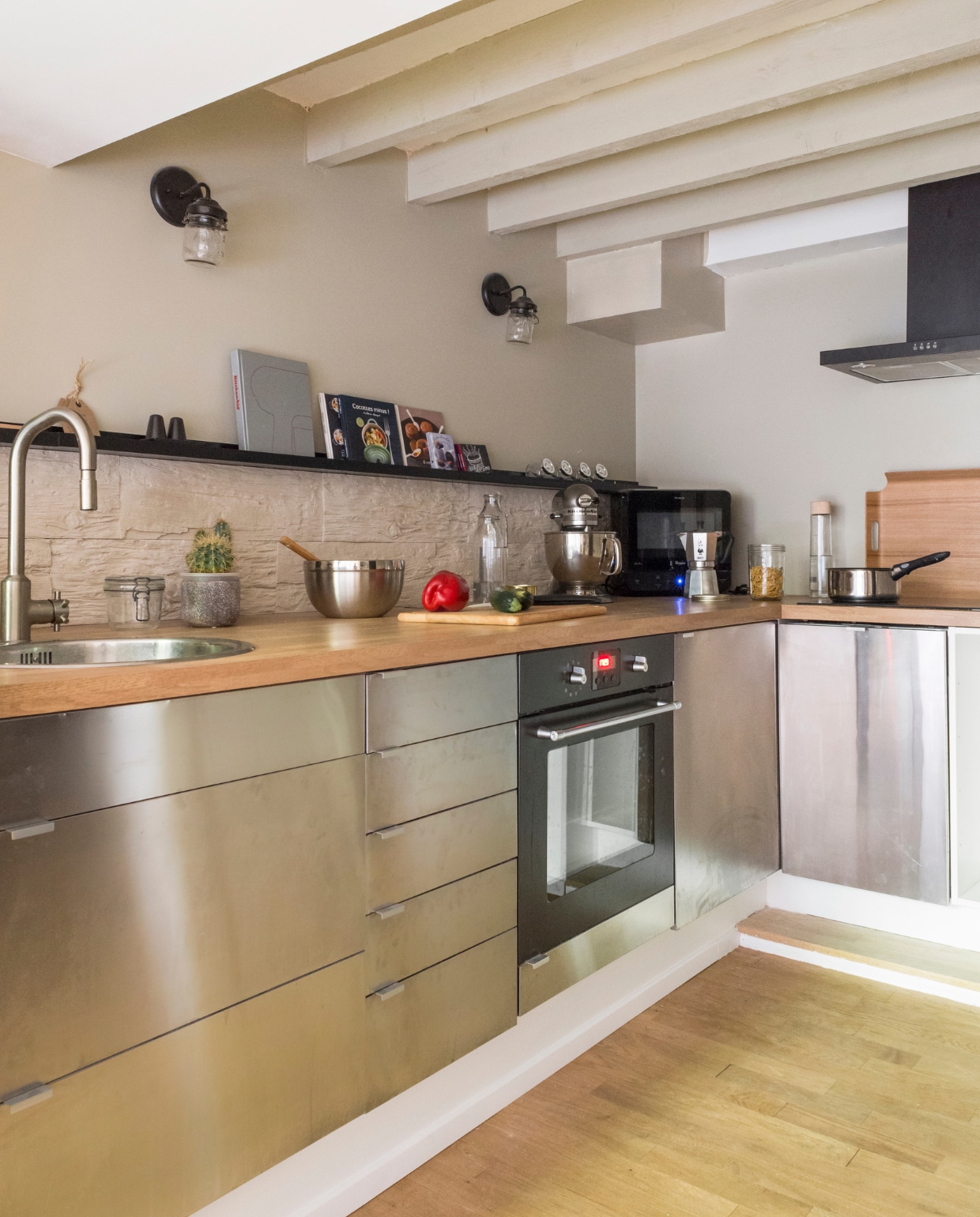 De keuken is van Ikea en werd uitgerust met een eikenhouten werkblad. Stenen panelen dienen als spatrand, afgezet met een donkere wandplank van Ikea. De wandlampen zijn gekocht op Etsy. Het trapje leidt naar de entree, langs een wand vol familiefoto’s, tekeningen en sierobjecten. Houten dienblad van Habitat, keukenmachine van Kitchen Aid. Bron: vtwonen 03-2015 | fotografie Julien Fernandez, amandinejules.com | tekst Amandine Berthon | tekstbewerking Monique van der Pauw