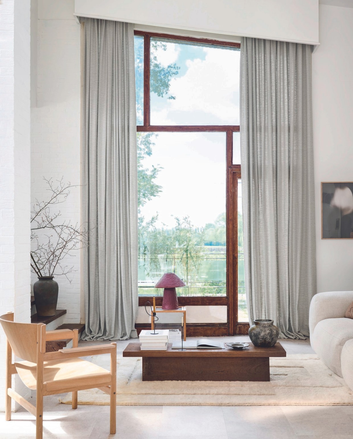 Lichtdoorlatend gordijn op maat in stof Blurred Lines, kleur Soft Grey (2322), per meter vanaf € 31,99. M.m.v. comfortmeubel.be (bank), noyastudio.dk (vloerkleed), moduleo.com (vloer), vtwonen.nl/shop (vitrinebox en kandelaar van Ferm Living), zarahome.com (tafellamp, kussen, bord), clairehorsten.com (hoge vaas), taktcph.com (fauteuil), eclatcreation.be (lage vaas). vtwonen 08-2024 | styling Kim van Rossenberg | fotografie Sjoerd Eickmans