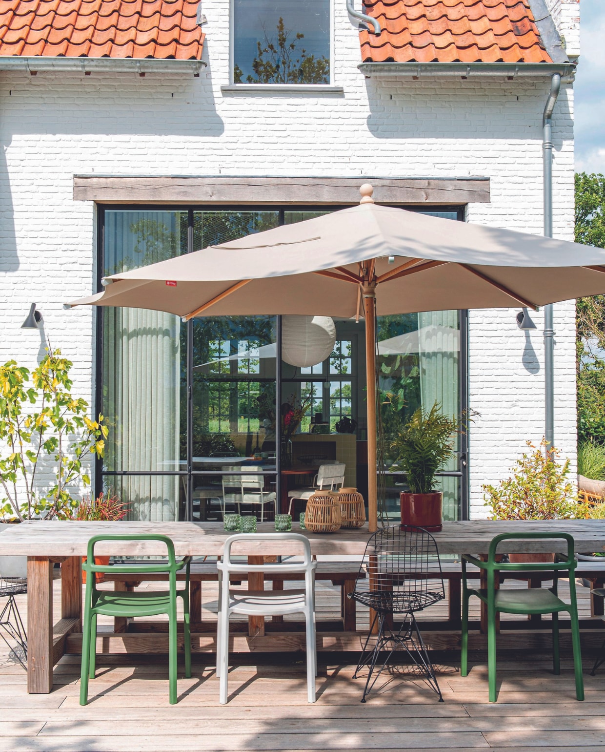 Houten tuintafel Marktplaats, groene en witte stoelen Ikea, zwarte stoelen Vitra, parasol Kees Smit. Waxinelichthouders en lantaarns Intratuin, wandlampen Louis Poulsen. vtwonen 08-2024 | styling Jeske Weel | fotografie Caroline Coehorst