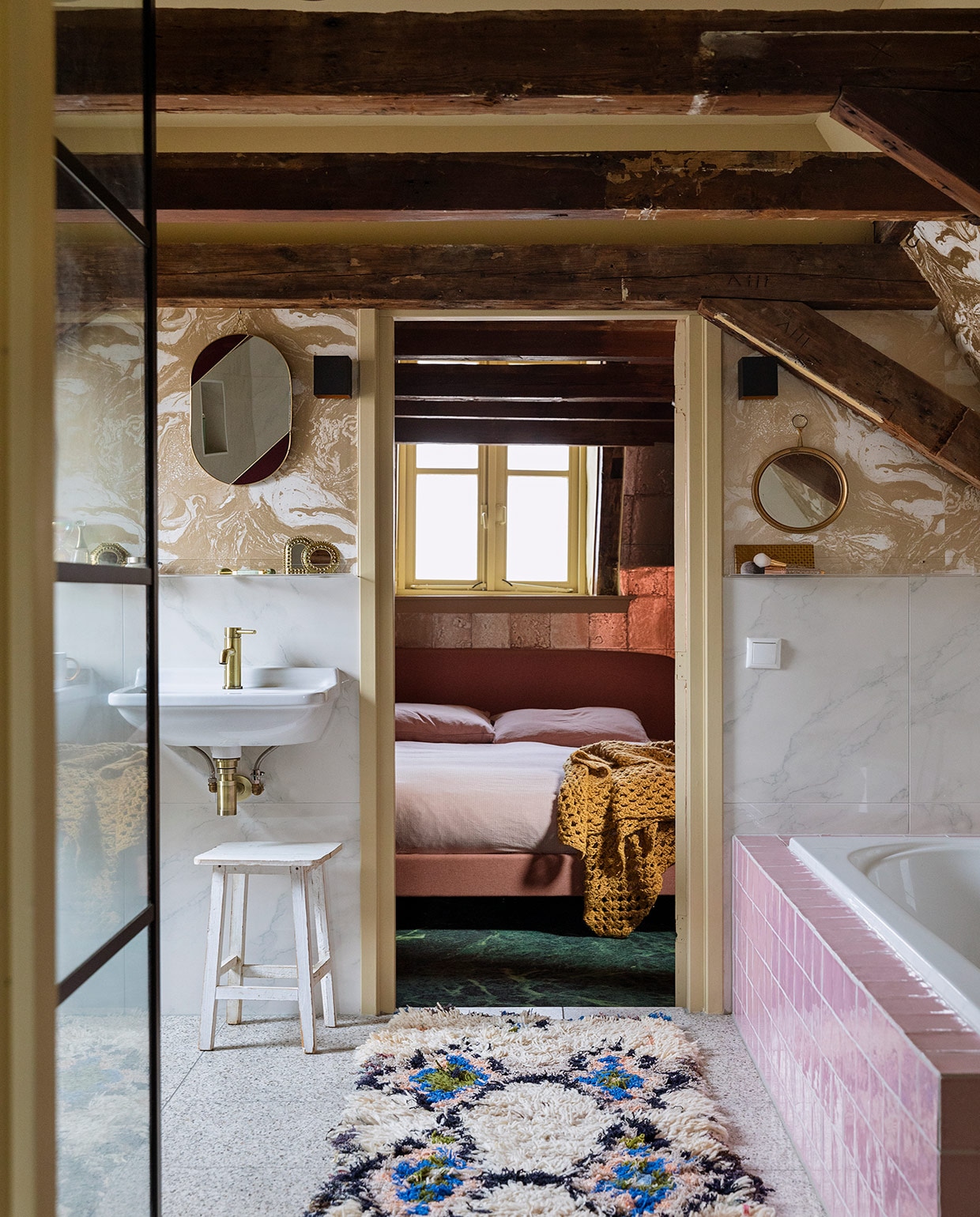 Terrazzo vloertegels, keramische wandtegels met marmerprint, kraan van geborsteld koper, roze badtegels en witte kruk herkomst onbekend. Behang Marble Gold 188 van Ferm Living. Wastafels Villeroy &amp; Boch. Ovalen spiegel &amp;Klevering. Ronde spiegel antiek. Kleed The Souks. In de slaapkamer: behang Antique Mirror in goud van Cole &amp; Son. Bed Made. Beddengoed Ikea. Sprei zelf gehaakt. vtwonen 06-2021 | styling Ilona Jongepier | fotografie Dana van Leeuwen