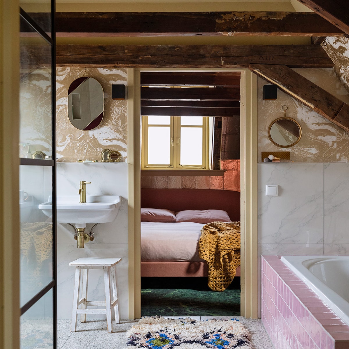 Terrazzo vloertegels, keramische wandtegels met marmerprint, kraan van geborsteld koper, roze badtegels en witte kruk herkomst onbekend. Behang Marble Gold 188 van Ferm Living. Wastafels Villeroy & Boch. Ovalen spiegel &Klevering. Ronde spiegel antiek. Kleed The Souks. In de slaapkamer: behang Antique Mirror in goud van Cole & Son. Bed Made. Beddengoed Ikea. Sprei zelf gehaakt.