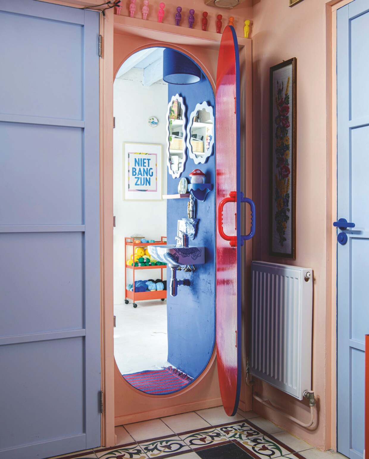 Muurverf Perzik en Retro Blauw van Decoverf, muurverf Cobalt Blue 2 van Lacq, verf op de deur Jachtrood van Karwei. Poppetjes Meneer Paprika, vloerkleed Hay, kast Ikea, poster Grafische Werkplaats Amsterdam, wasbak Tattootoilet Esther Derkx, paddenstoellamp It’s a present, wandplank Studio Harm en Elke, spiegels Xenos. Vloertegels lagen al in het huis. Ovale deur zelf ontworpen en laten maken, deurgrepen Bol, deurbeslag op rechter deur van Colombo Design via Schreuder &amp; Co. vtwonen 09-2024 | styling Inge van Lieshout | fotografie Louis Lemaire