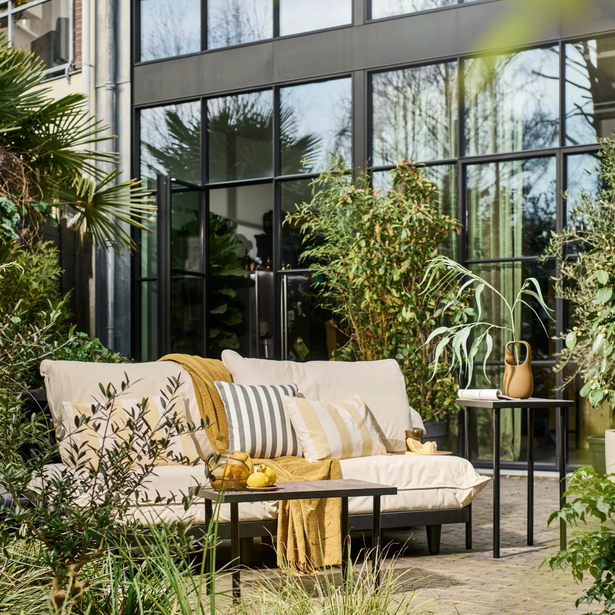 Modulaire loungeset Paletti van Fatboy, kunststof frame met waterafstotende kussens € 899,-, bruine stenen vaas Nicita van Bloomingville € 58,90 (vtwonen.nl/shop). Plaid € 12,99, blauw gestreept kussen € 17,99, geel gestreept kussen € 17,99, glazen karaf € 29,99, houten onderzetter € 5,99, houten dienblad € 17,99, drinkglazen € 5,99 (zarahome.com). Overig privébezit. vtwonen 08-2023 | styling en uitvoering Sanne Ketelaar | fotografie Sjoerd Eickmans