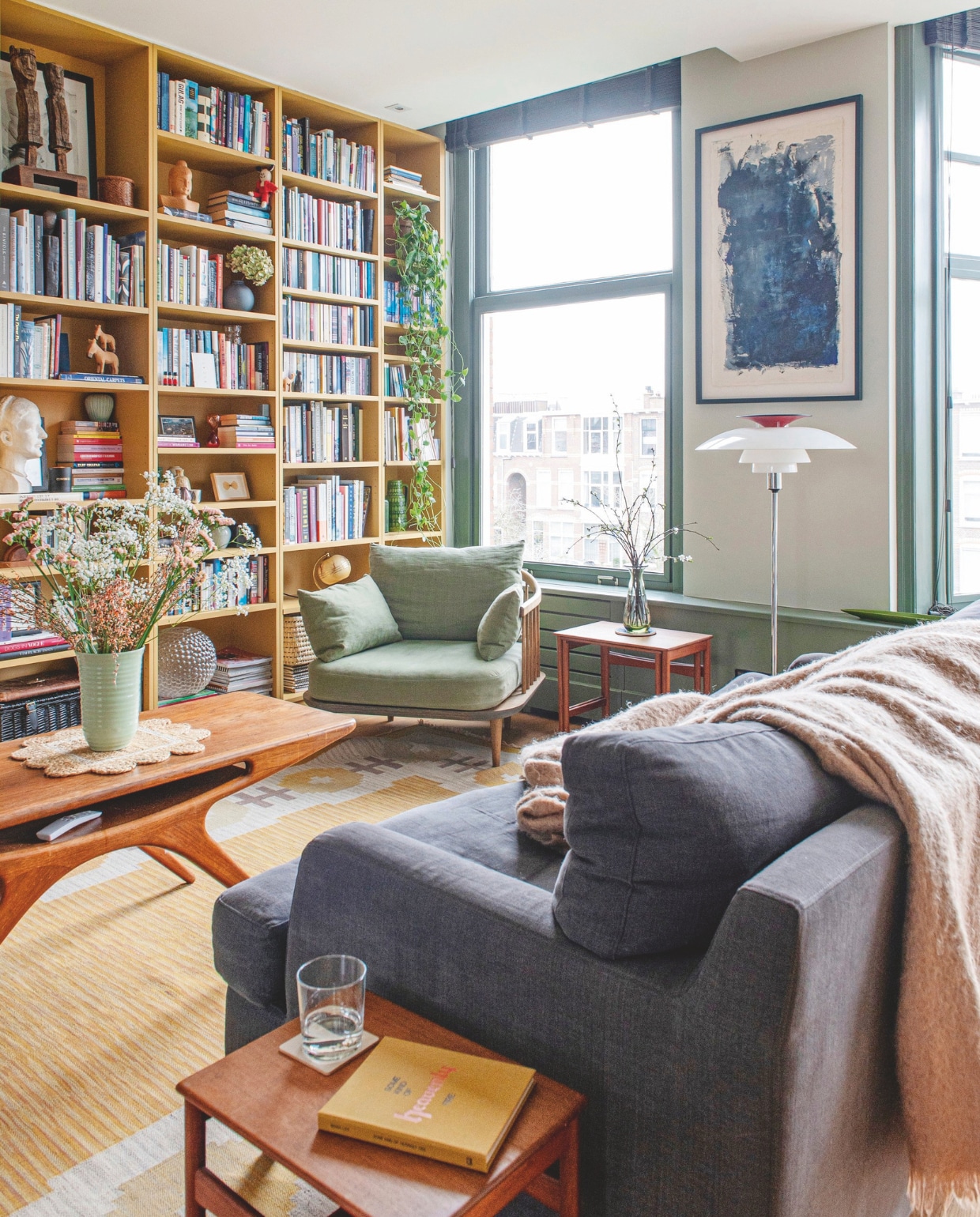 Boekenkast maatwerk, geverfd in India Yellow van Farrow &amp; Ball, fauteuil Fly Chair SC1 &amp;Tradition, vintage koffietafel Smile Johannes Andersen, Deense vintage bijzettafels, staande lamp PH80 Poul Henningsen, kunstwerk van Victoria Collinge, bank Sofa.com, mohair plaid By Mölle, vintage vaas Denby, placemat Sunday &amp; Story, karpet We Are Here Now, kleur raamkozijnen Green Smoke van Farrow &amp; Ball. vtwonen 11-2024 | stying Jeske Weel | fotografie Caroline Coehorst